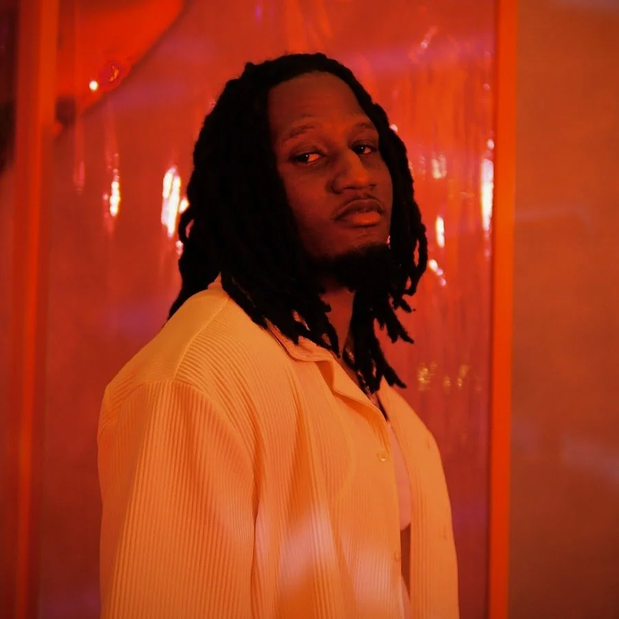 A man with dreadlocks and a slight beard, wearing a yellow/orange shirt, standing in front of a reflective red background.