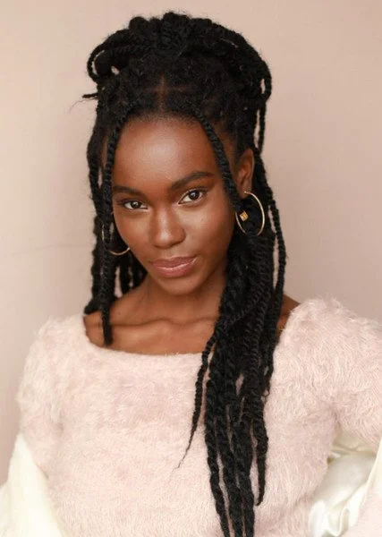 A woman with long, black dreadlocks wearing a light pink fuzzy sweater and gold hoop earrings.