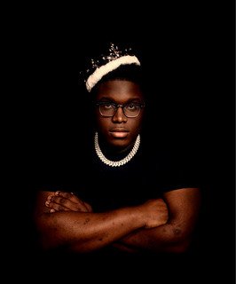 Person wearing glasses, a pearl necklace, and a tiara, with a black background.