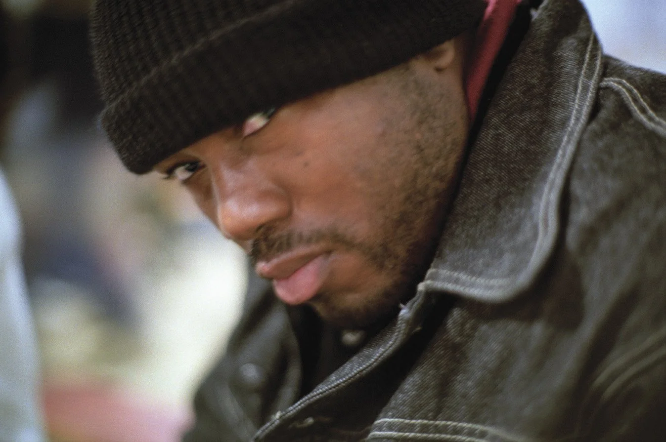 Close-up of a man wearing a dark beanie and a denim jacket, looking to the side with a serious expression.