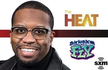 Close-up of a smiling man wearing glasses, with logos for 'The Heat', 'SiriusXM FLY', and 'SXM' in the background.