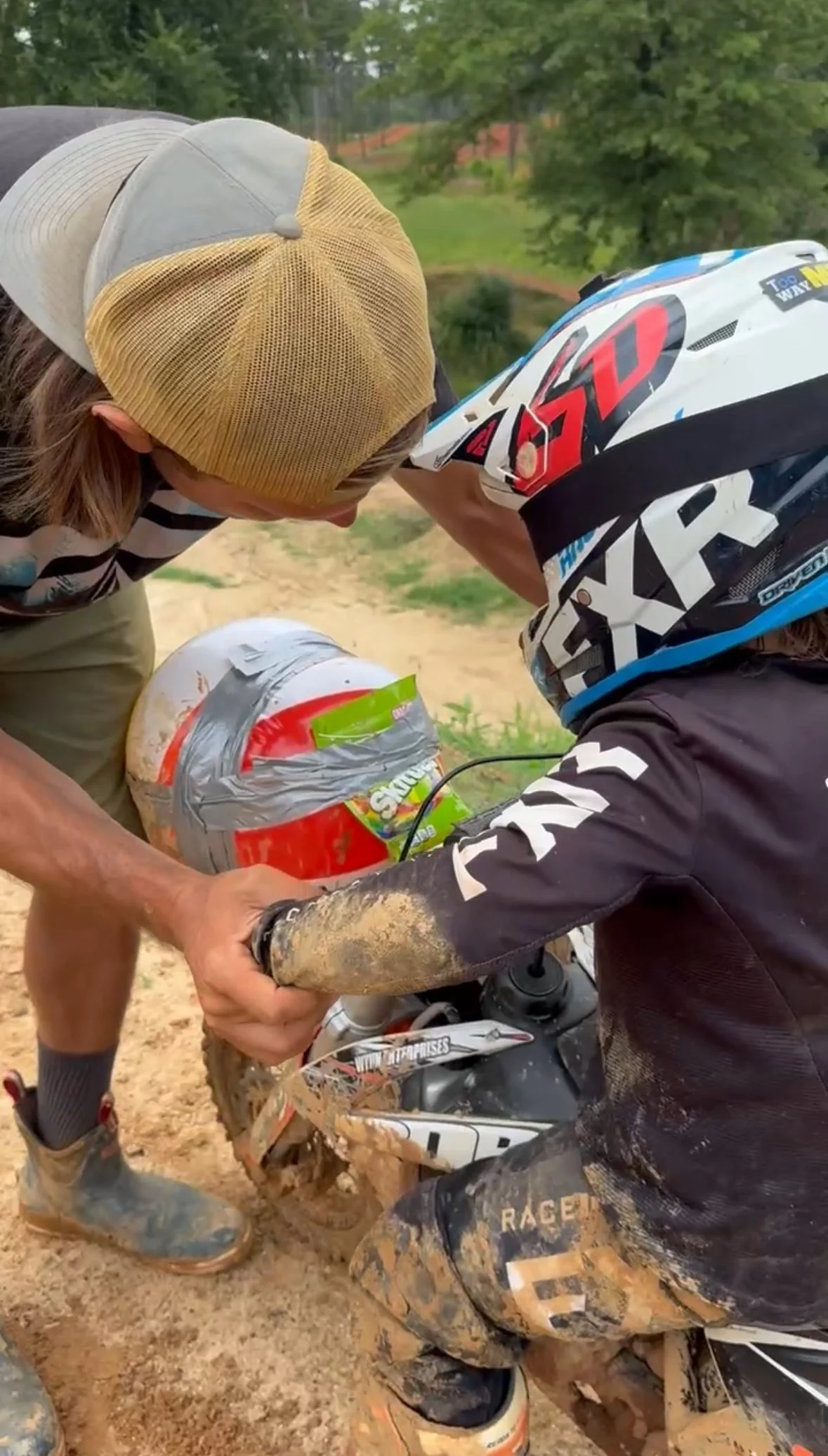 A person helping a young dirt bike rider check their helmet and bike after a muddy ride outdoors.