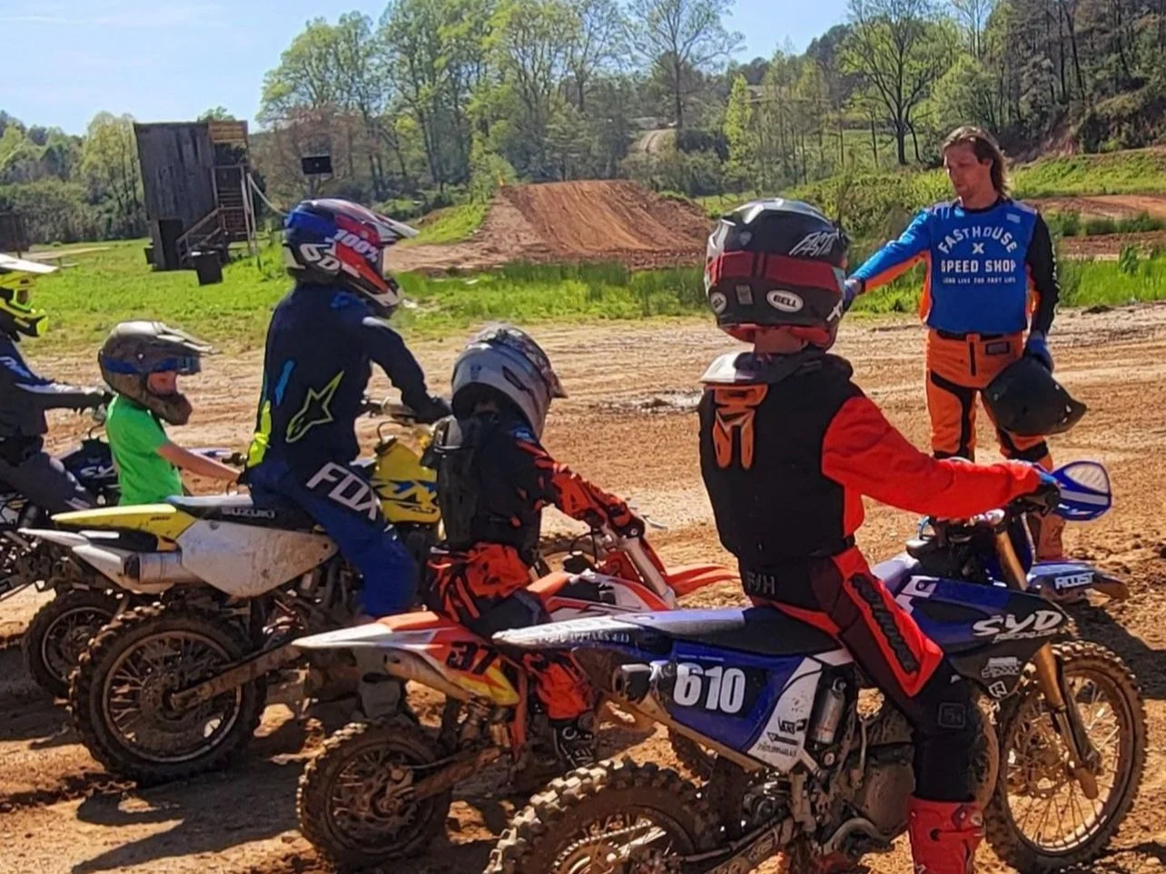 Group of children and an adult on motocross bikes at a dirt track, with dirt jumps and green trees in the background, sunny day.