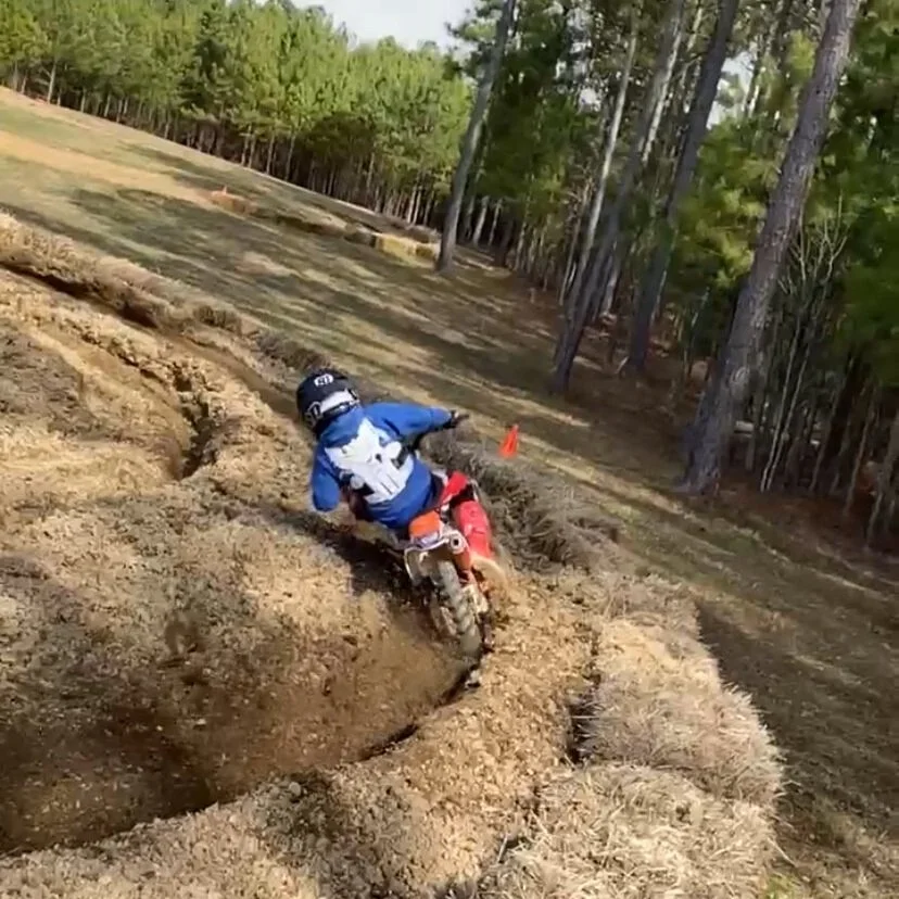 A person riding a dirt bike through a track in a forested area with trees and clear sky.