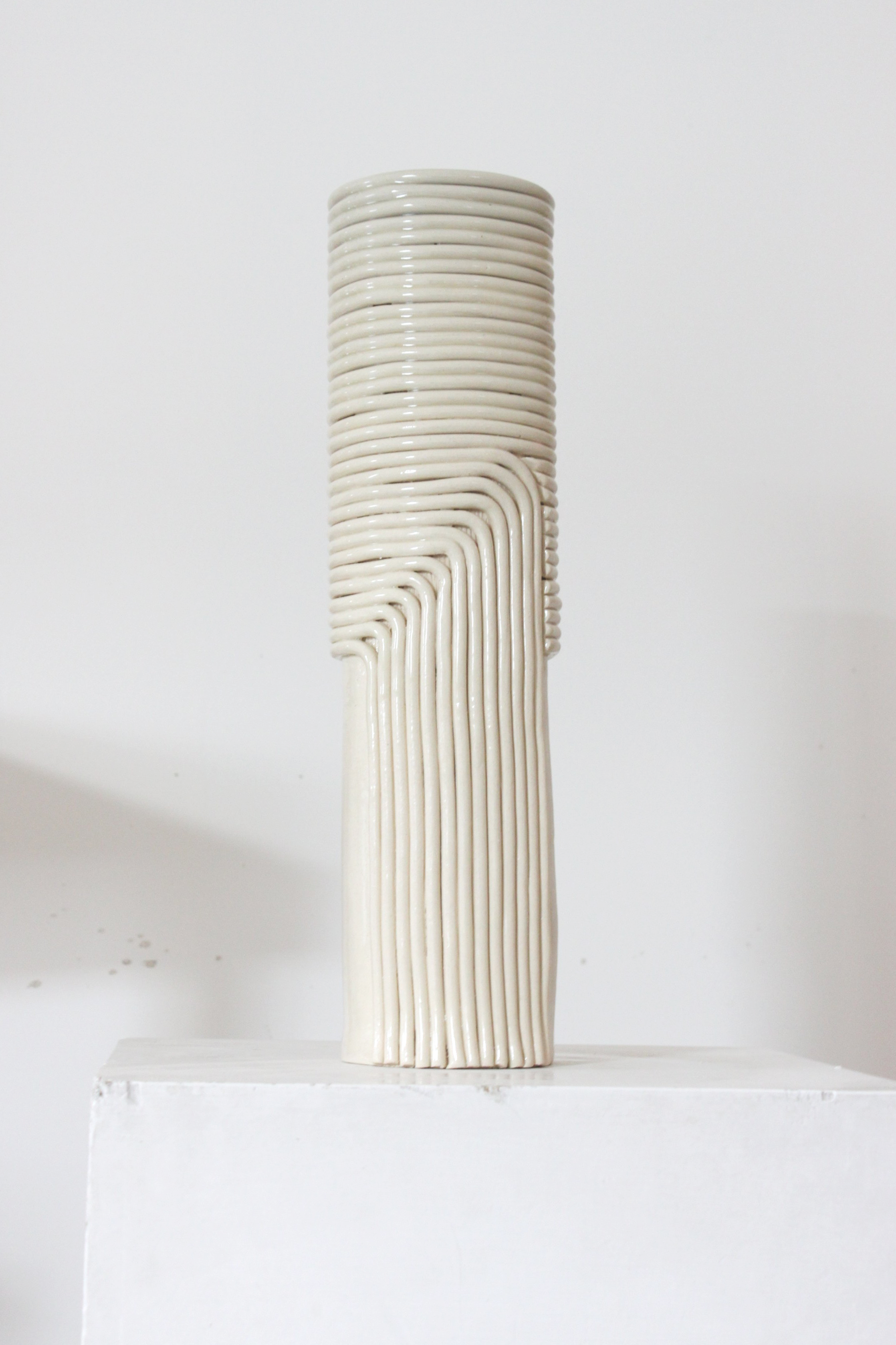 Une sculpture en céramique blanche avec une forme cylindrique composée de lignes incurvées, placée sur un socle blanc.