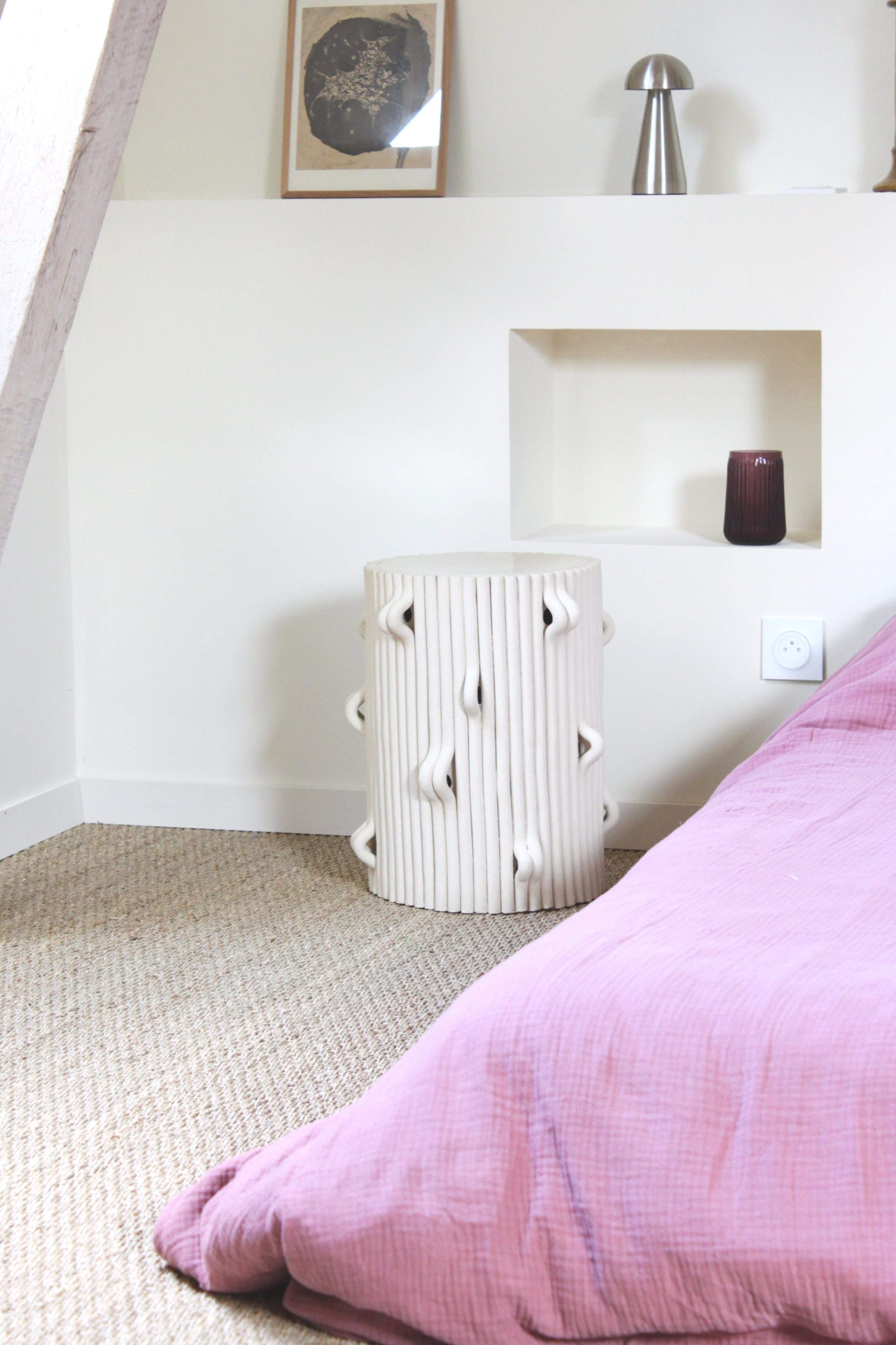 Une pièce avec un fauteuil blanc sculptural, un coussin rose en tissu, un mur blanc avec une niche contenant une vase sombre, et une lampe en métal. Il y a également un cadre d'art au mur.