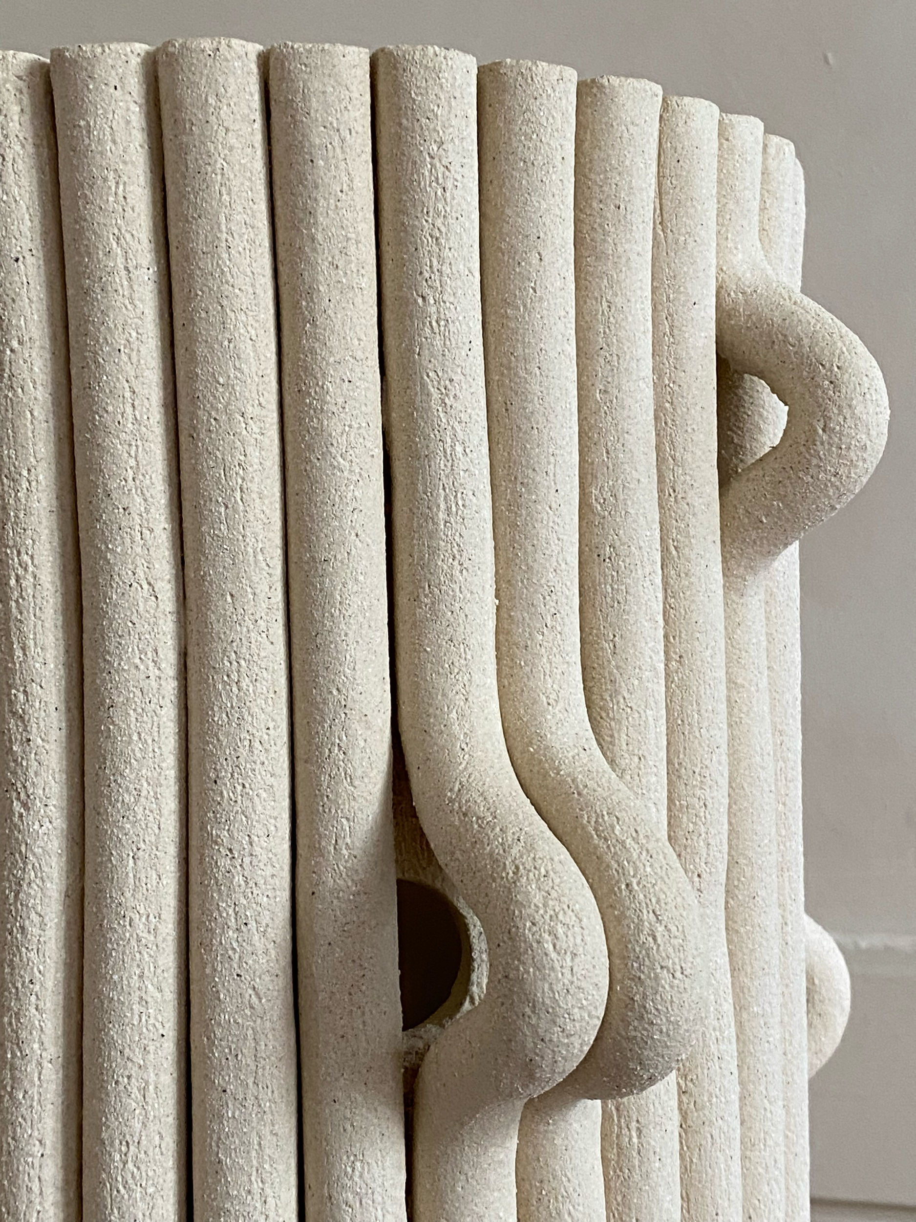 Une sculpture en forme de mug créée à partir de tubes en céramique ou en terre cuite de couleur beige clair, avec une poignée sur le côté droit.