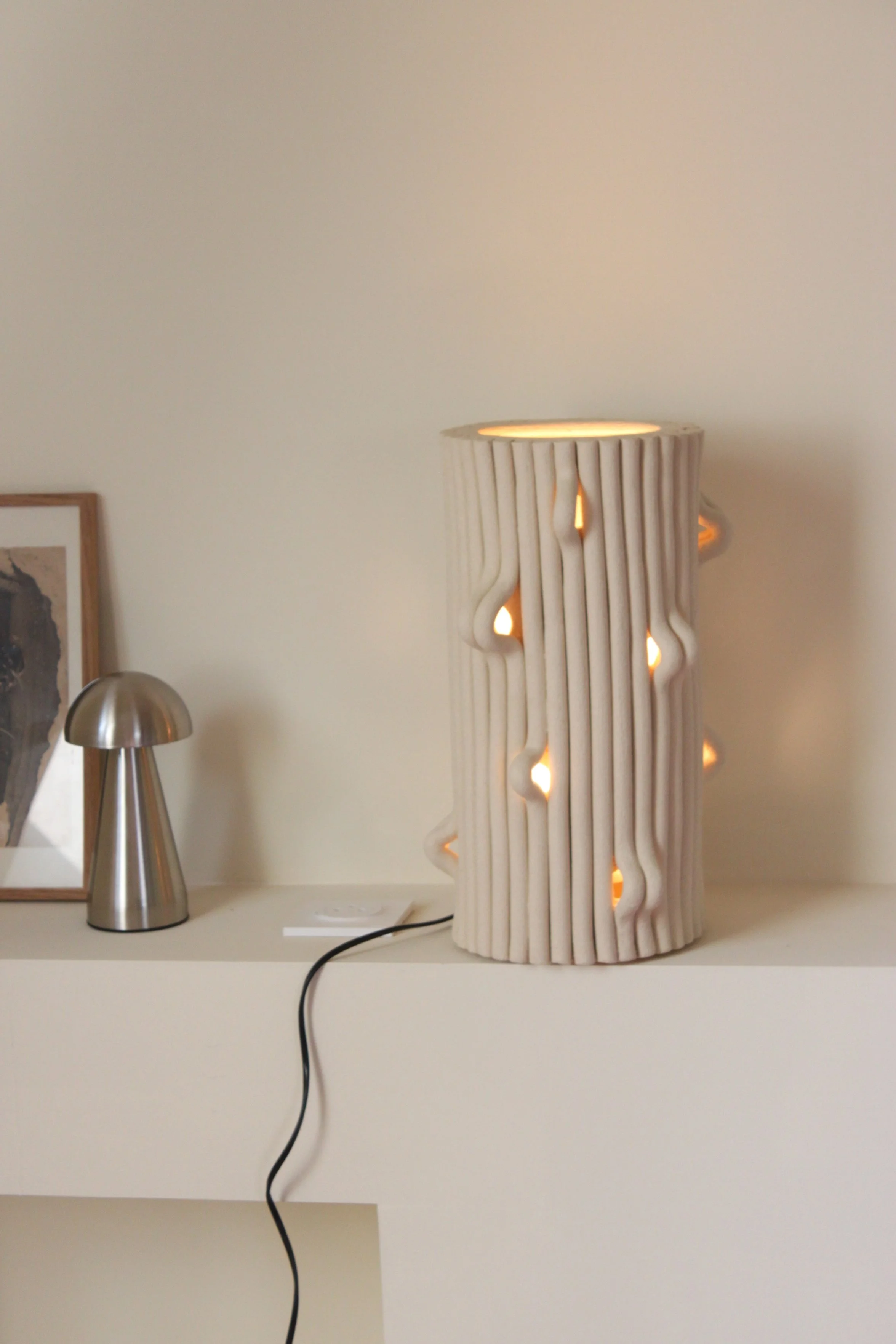 Lampe décorative en céramique blanche avec un motif ondulé et des lumières chaudes à l'intérieur, posée sur une étagère blanche.