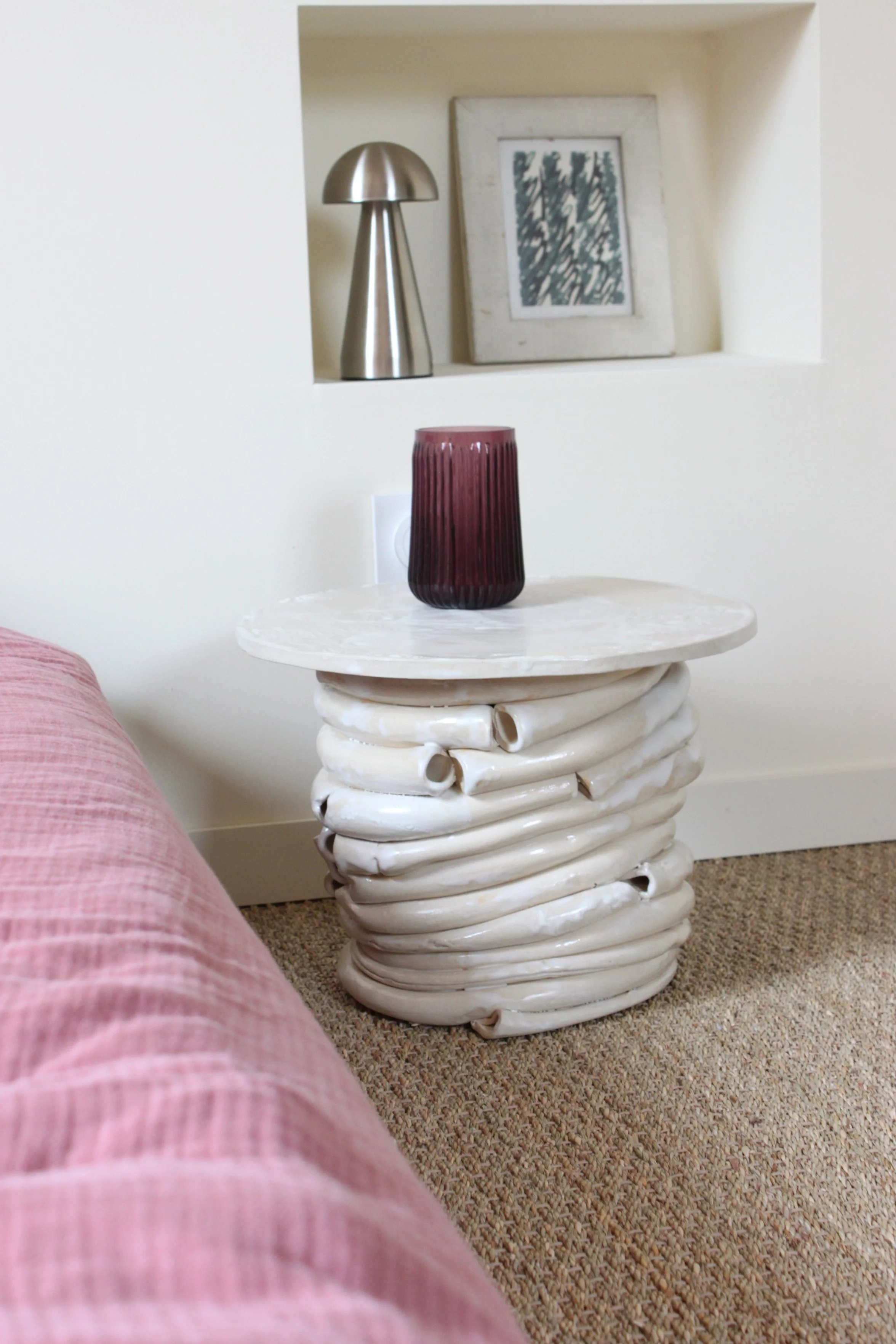 Table d'appoint en céramique blanche avec une base torsadée, surmontée d'une surface ronde, avec un vase violet foncé, en verre, dessus. En arrière-plan, un mur blanc avec une niche contenant une lampe en métal argenté et une œuvre d'art encadrée.