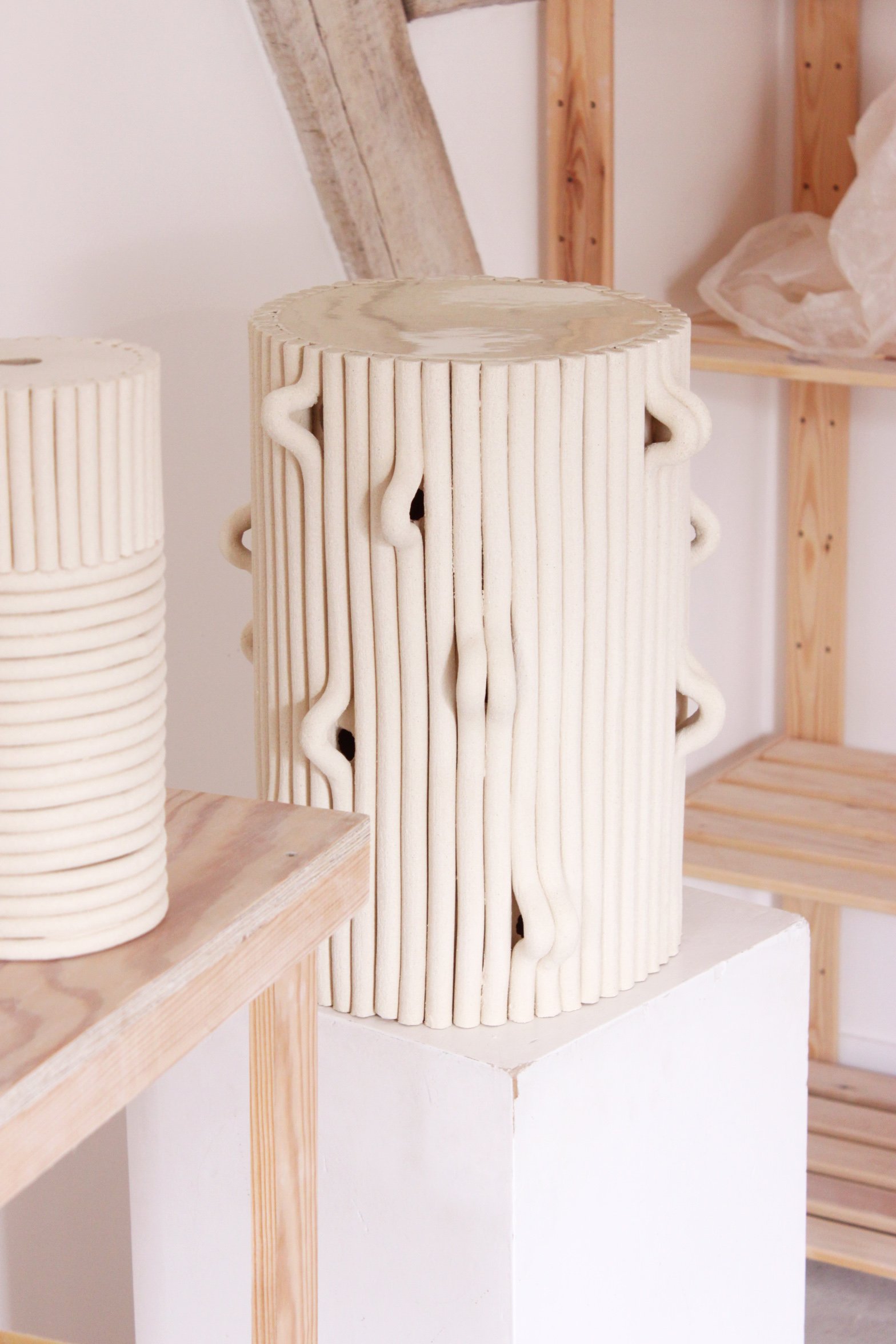 Une sculpture en céramique blanche cylindrique avec des motifs ondulés et des lignes verticales, exposée sur un socle blanc dans un espace d'exposition