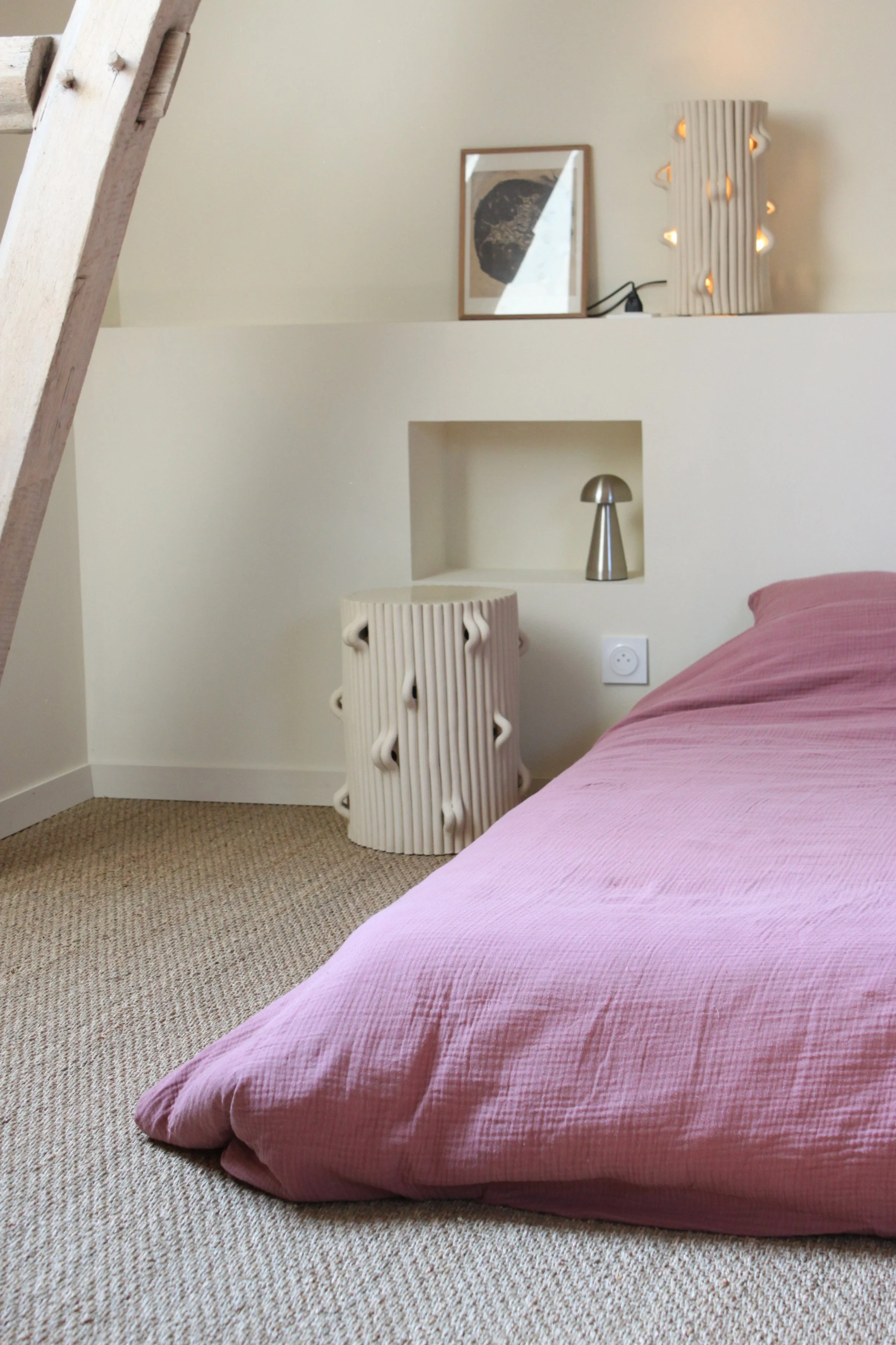 Une chambre avec un lit à linge rose, un tapis beige, une table d'appoint en céramique avec des motifs ondulés, une lampe de chevet en métal, et des décorations modernes sur un mur blanc avec une étagère