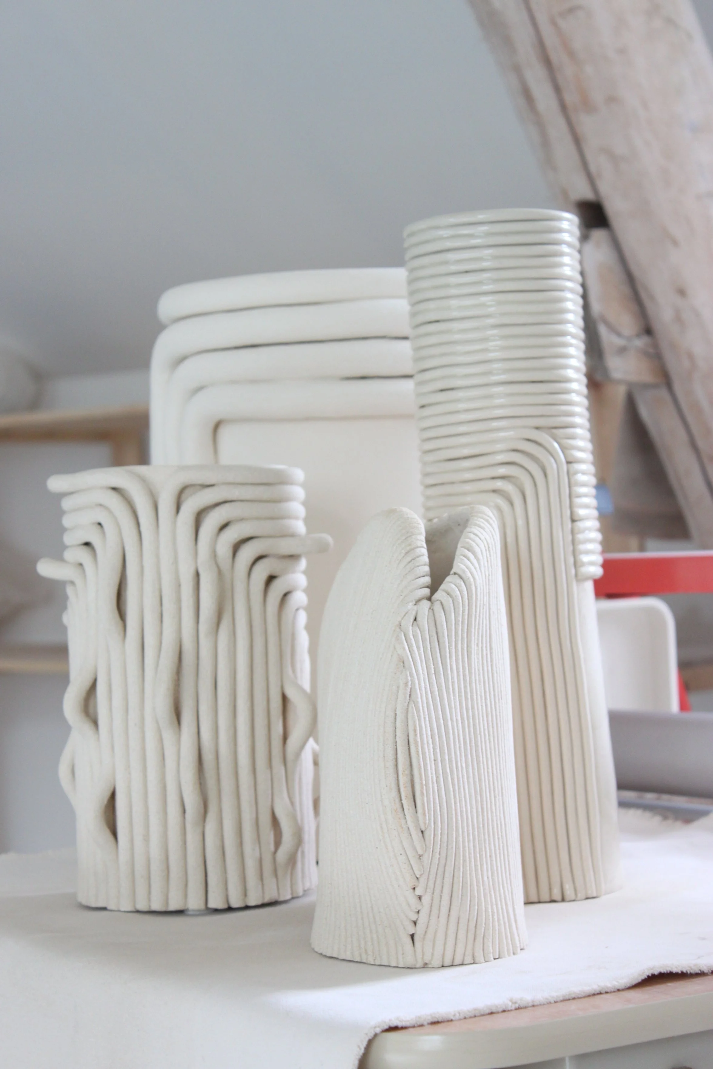 Piles de vaisselle en céramique blanche, comprenant des assiettes, des plats, des tasses et des bols, empilés sur une surface en tissu blanc dans un environnement intérieur.