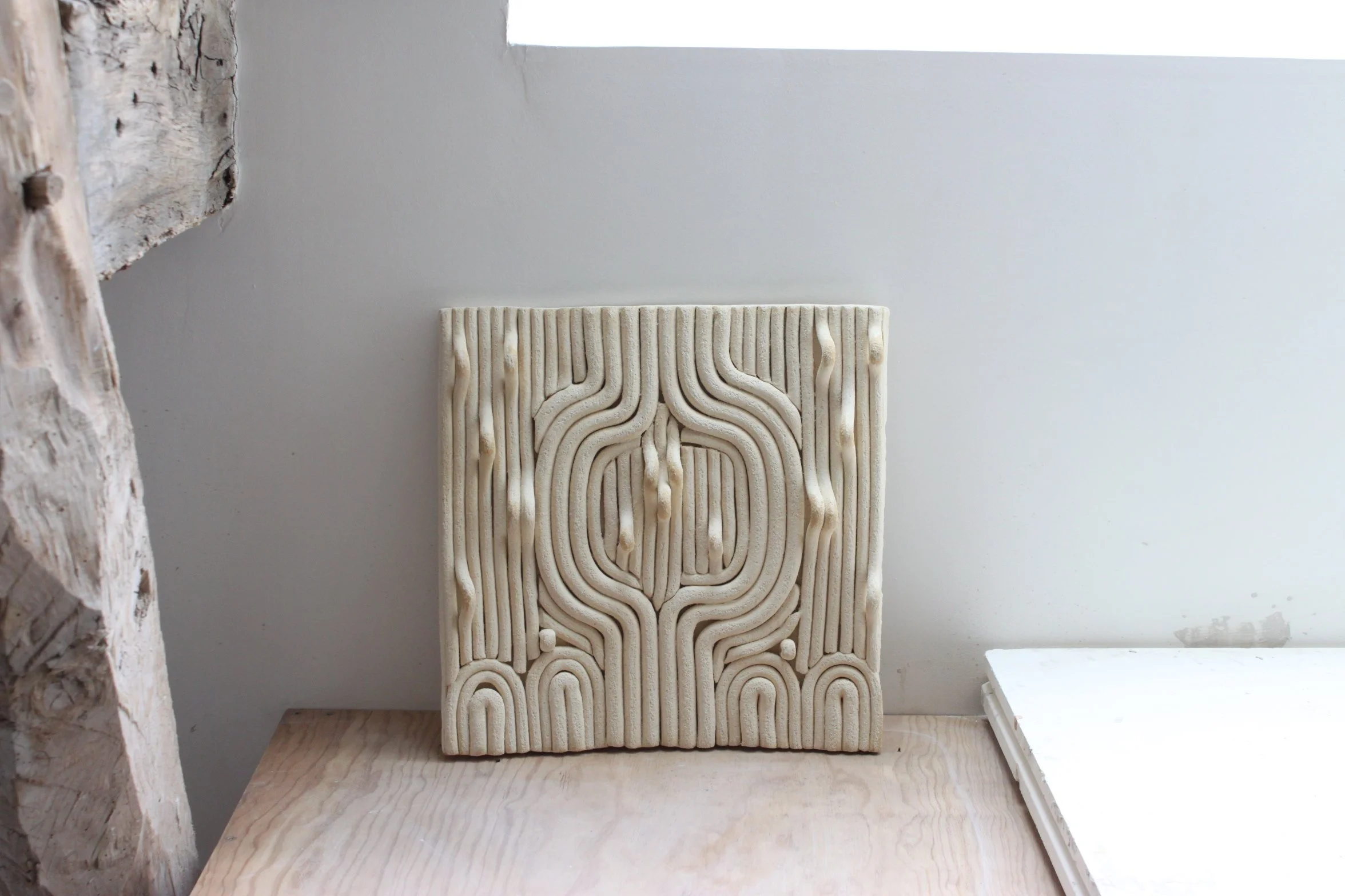 Une œuvre d'art murale dit bas-relief en grès blanc chamotte, en relief blanc avec un motif géométrique circulaire et rectiligne, posée sur une table en bois clair.