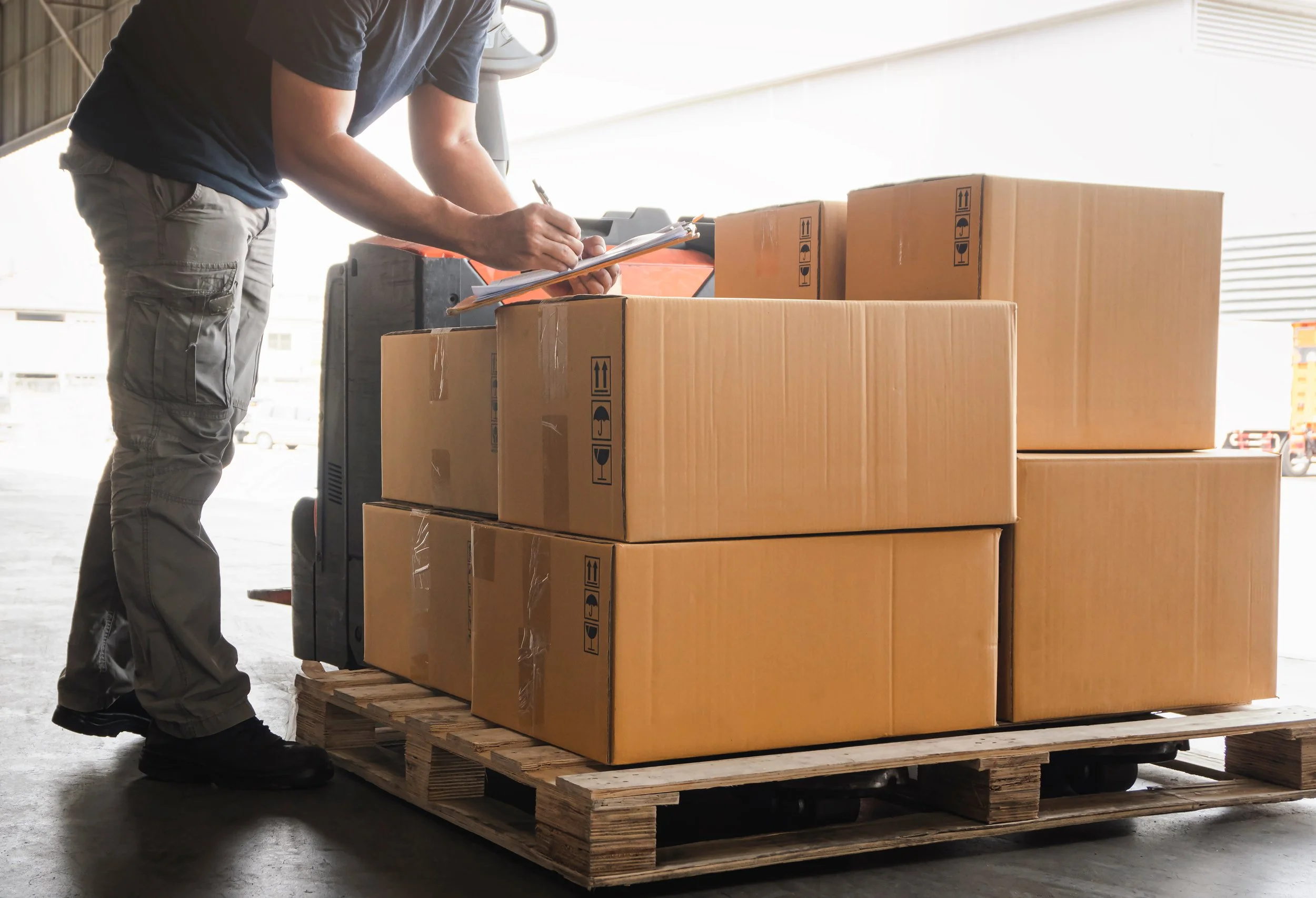 Person taking inventory on clipboard of boxes on a wooden pallet in a warehouse.