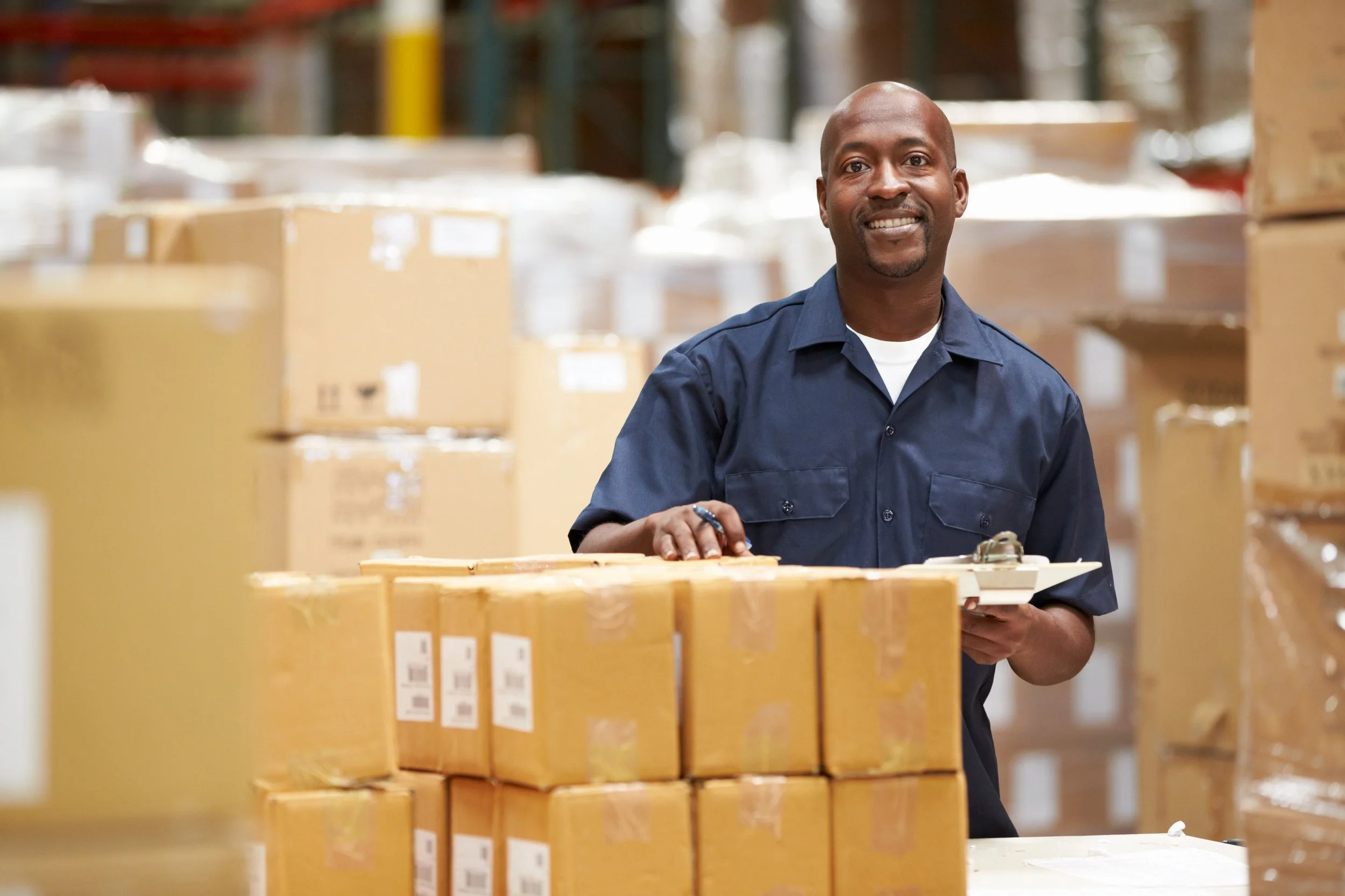 Man behind a stack of boxes smiling while holding an inventory clipboard.