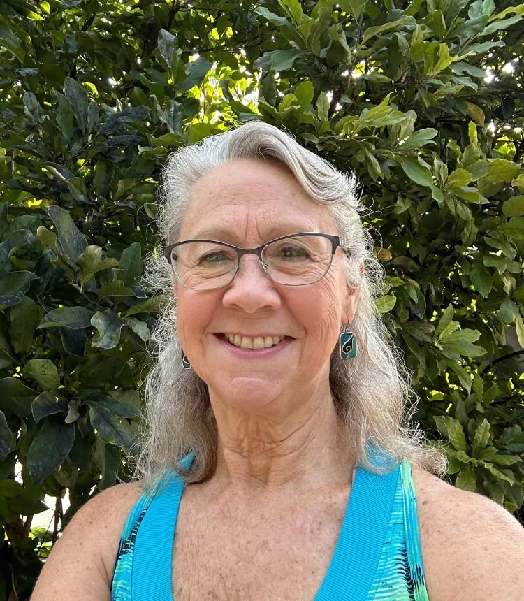 Older woman with gray hair, glasses, blue earrings, and a blue athletic top, smiling outdoors in front of green leafy bushes.