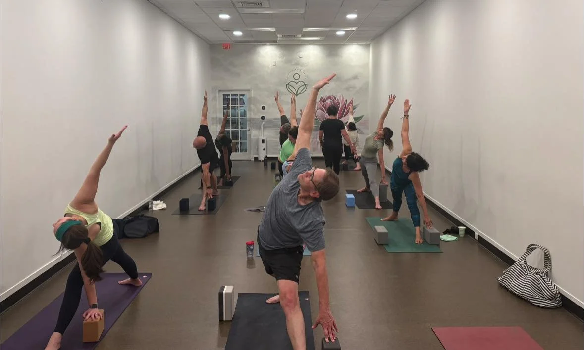 People practicing yoga in a spacious, well-lit room with white walls, some with mural art, on yoga mats with blocks and straps.
