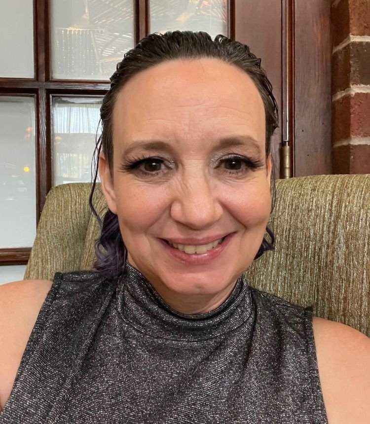 Close-up of a woman with dark hair and makeup, smiling while seated on a green-patterned armchair in a room with windows and wooden paneling.
