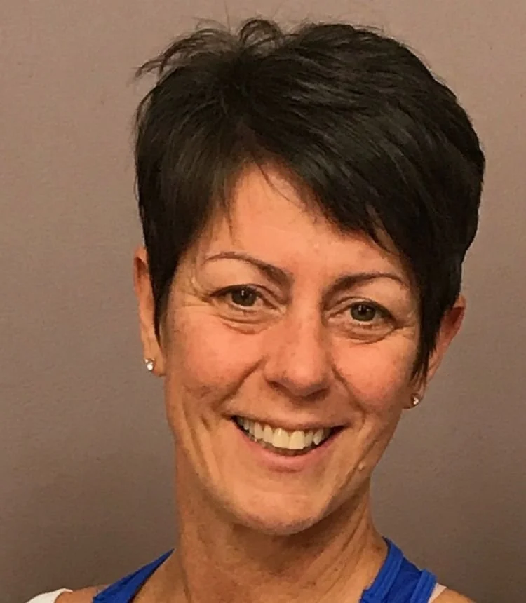 A woman with short dark hair smiling, wearing a white top and earrings, against a plain background.