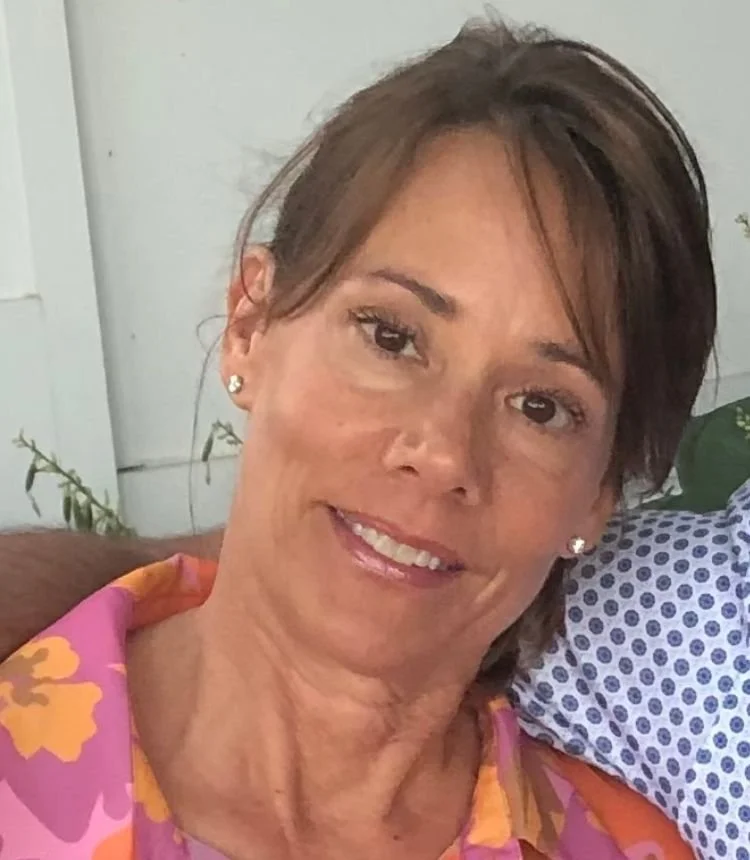 A woman with short brown hair and pearl earrings smiling, wearing a colorful pink, orange, and purple floral shirt.
