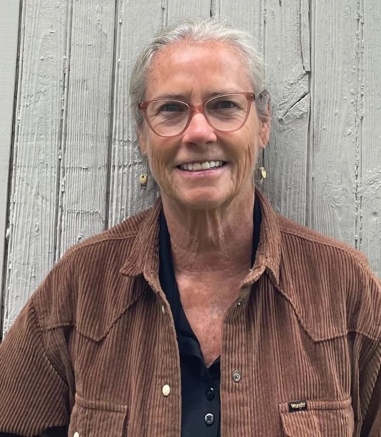 An older woman with gray hair, wearing glasses and earrings, smiles while standing outdoors in front of a gray, wooden fence, wearing a brown corduroy jacket and a black shirt.