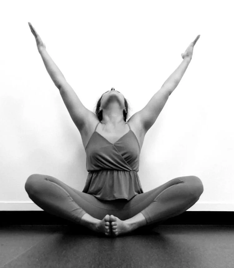 A woman practicing yoga or meditation, sitting cross-legged with arms raised upward and head tilted back, in a relaxed pose.