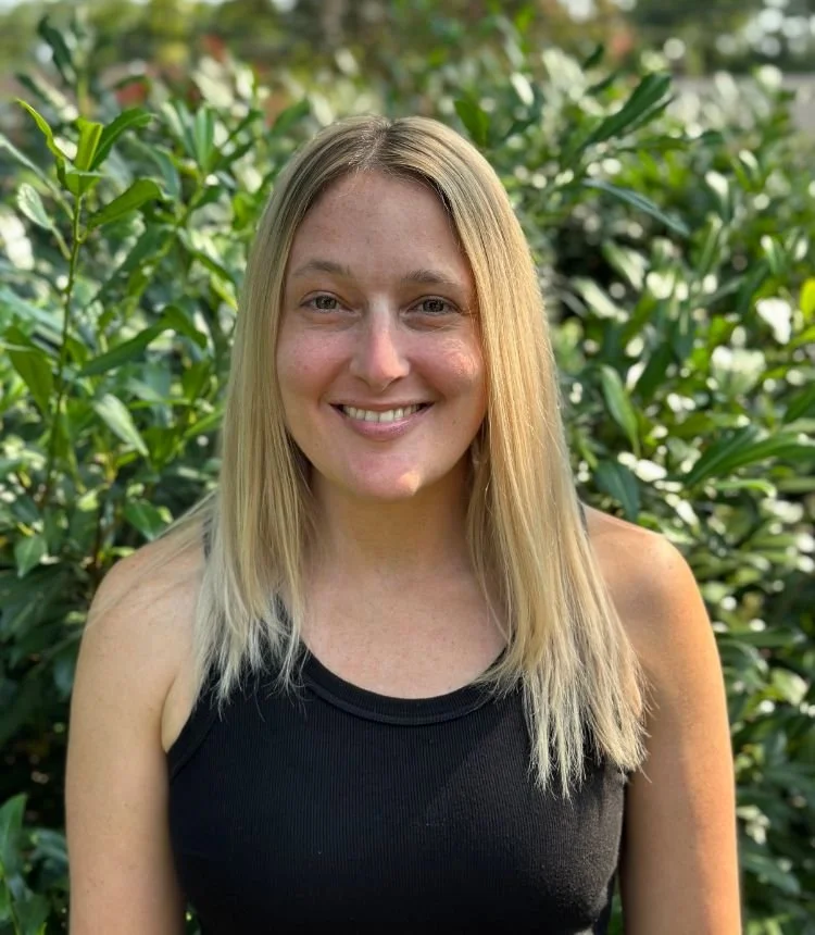 A young woman with blonde hair smiling outdoors in front of green bushes.