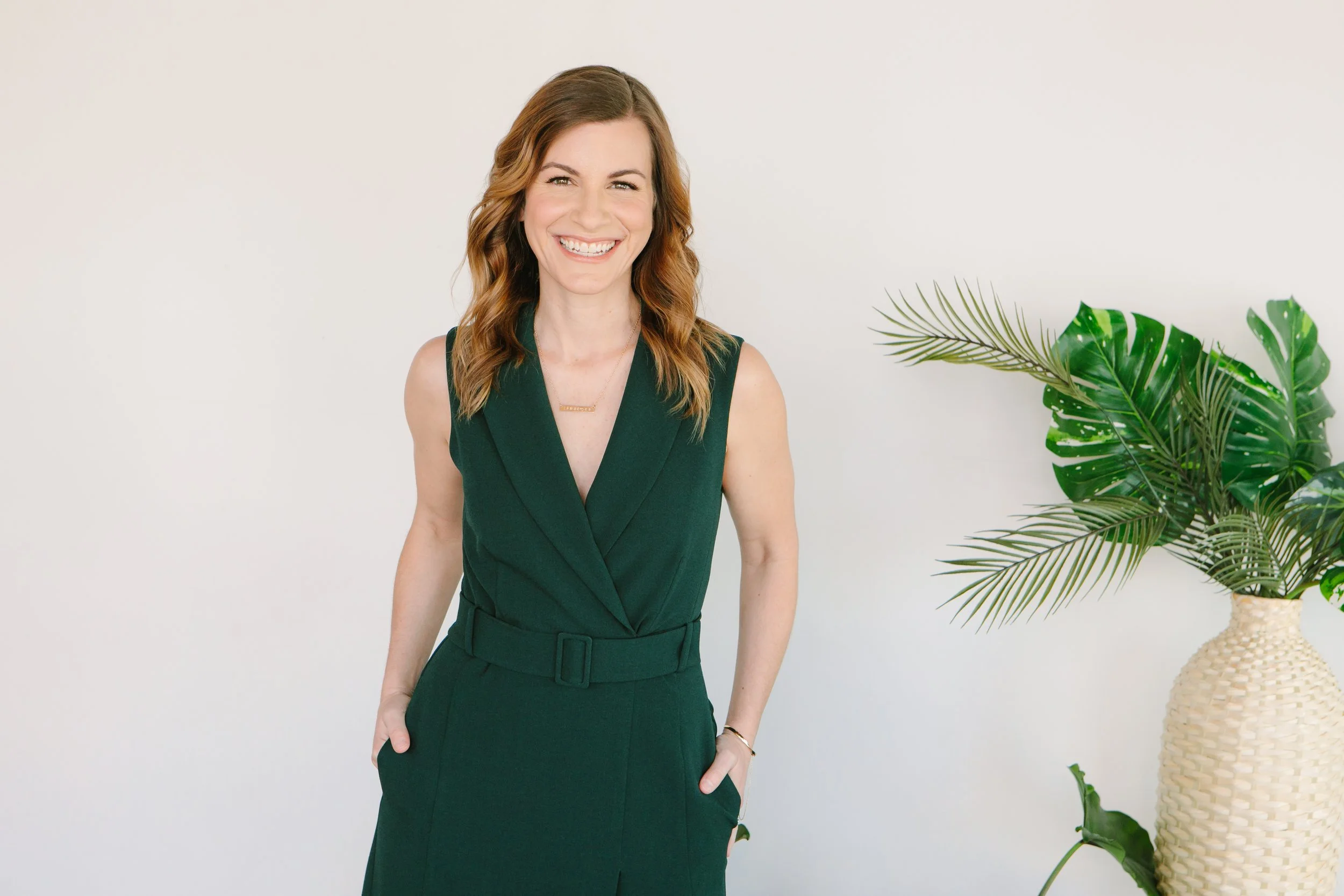 A woman with shoulder-length wavy brown hair and fair skin, wearing a sleeveless dark green dress with a belt, smiling with hands in pockets, standing near a large potted plant with broad green leaves in a light-colored woven vase against a plain white background.