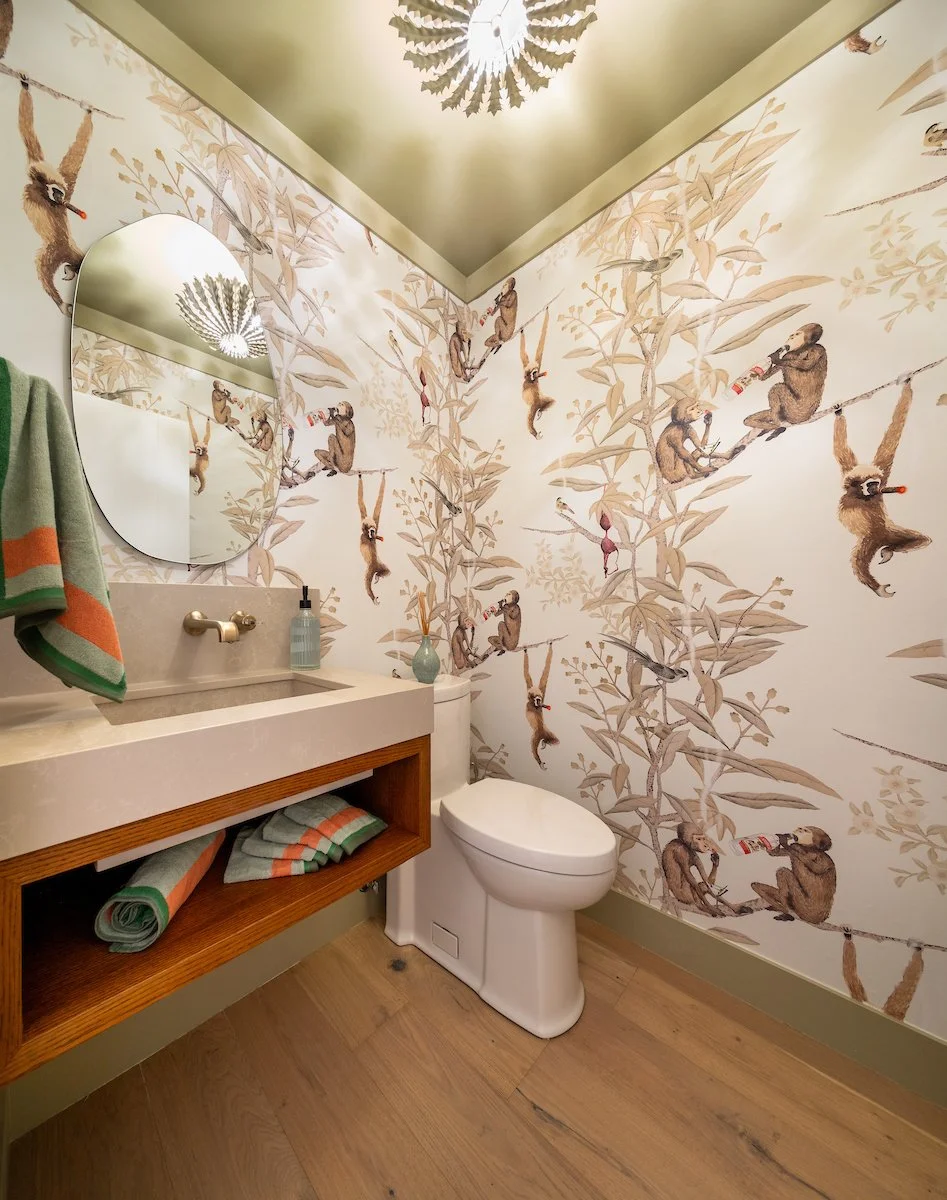 Bathroom with wallpaper featuring monkeys and bamboo, small sink with soap dispenser, towel with green, orange, and beige stripes, toilet, mirror, and wooden shelf with towels.