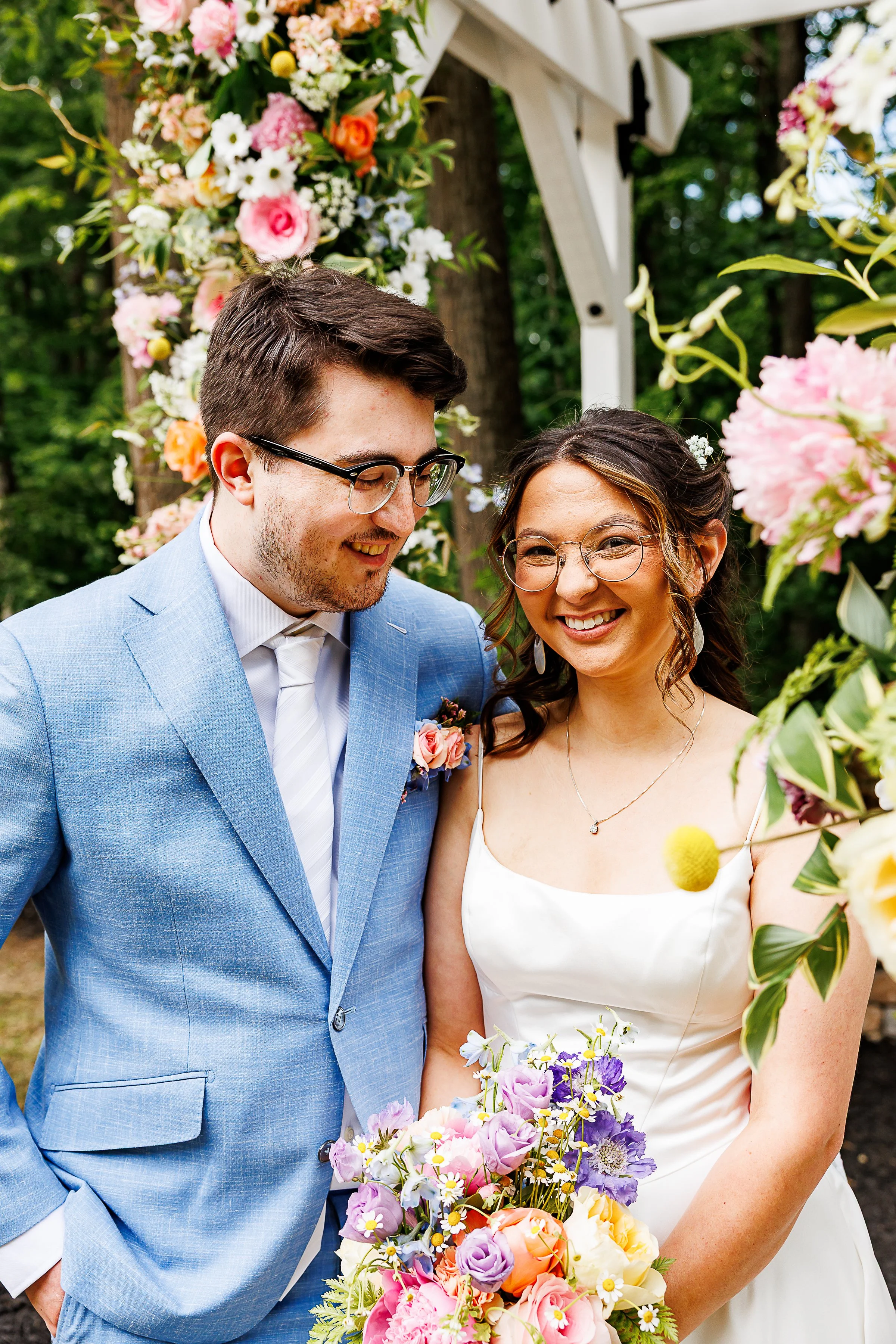 Bride and groom smiling together beneath a colorful floral arch during an outdoor wedding ceremony, with the bride holding a bright spring bouquet and the groom wearing a light blue suit.