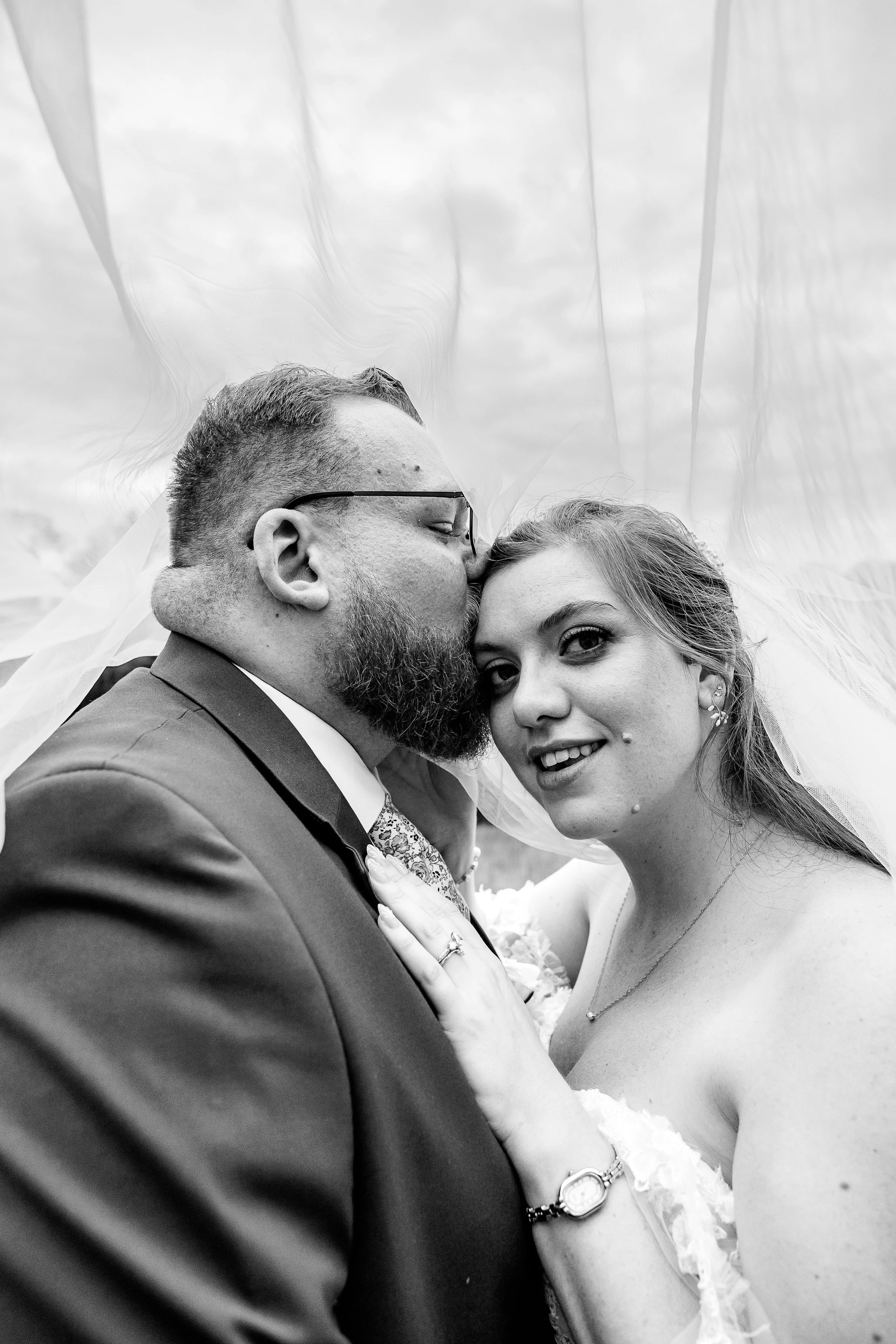 A tender black-and-white portrait of a newlywed couple beneath the bride’s veil, softly draped around them. The groom leans in to kiss her forehead while she smiles gently toward the camera, her hand resting on his chest, capturing an intimate moment