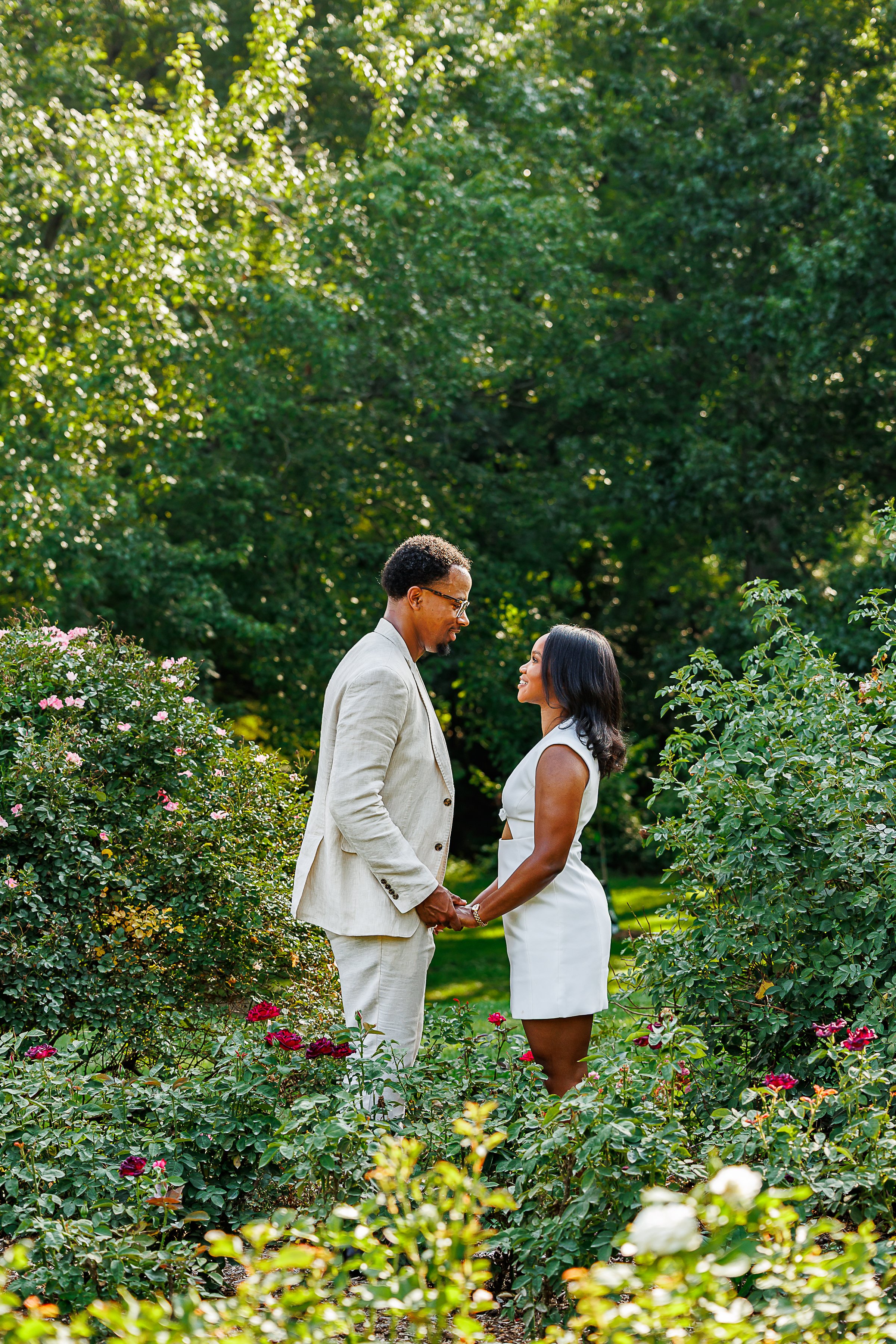 A couple stands hand in hand among blooming rose bushes in a vibrant garden. Facing one another with warm smiles, they wear coordinated light-colored outfits as dense greenery and colorful flowers frame the peaceful outdoor scene.