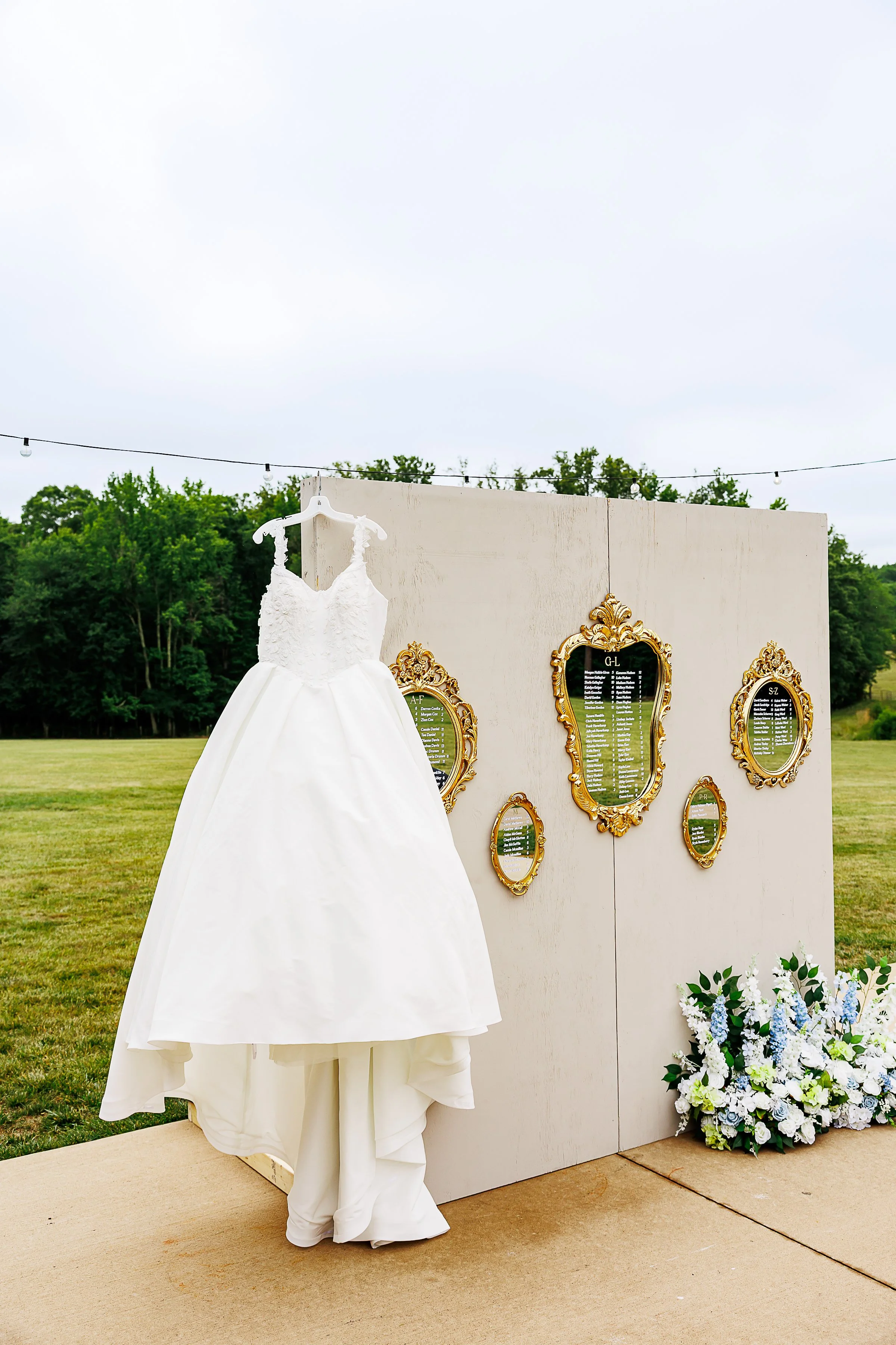 A white wedding dress displayed on a mannequin outdoors with a backdrop of firework displays and seating charts, beneath cloudy skies.