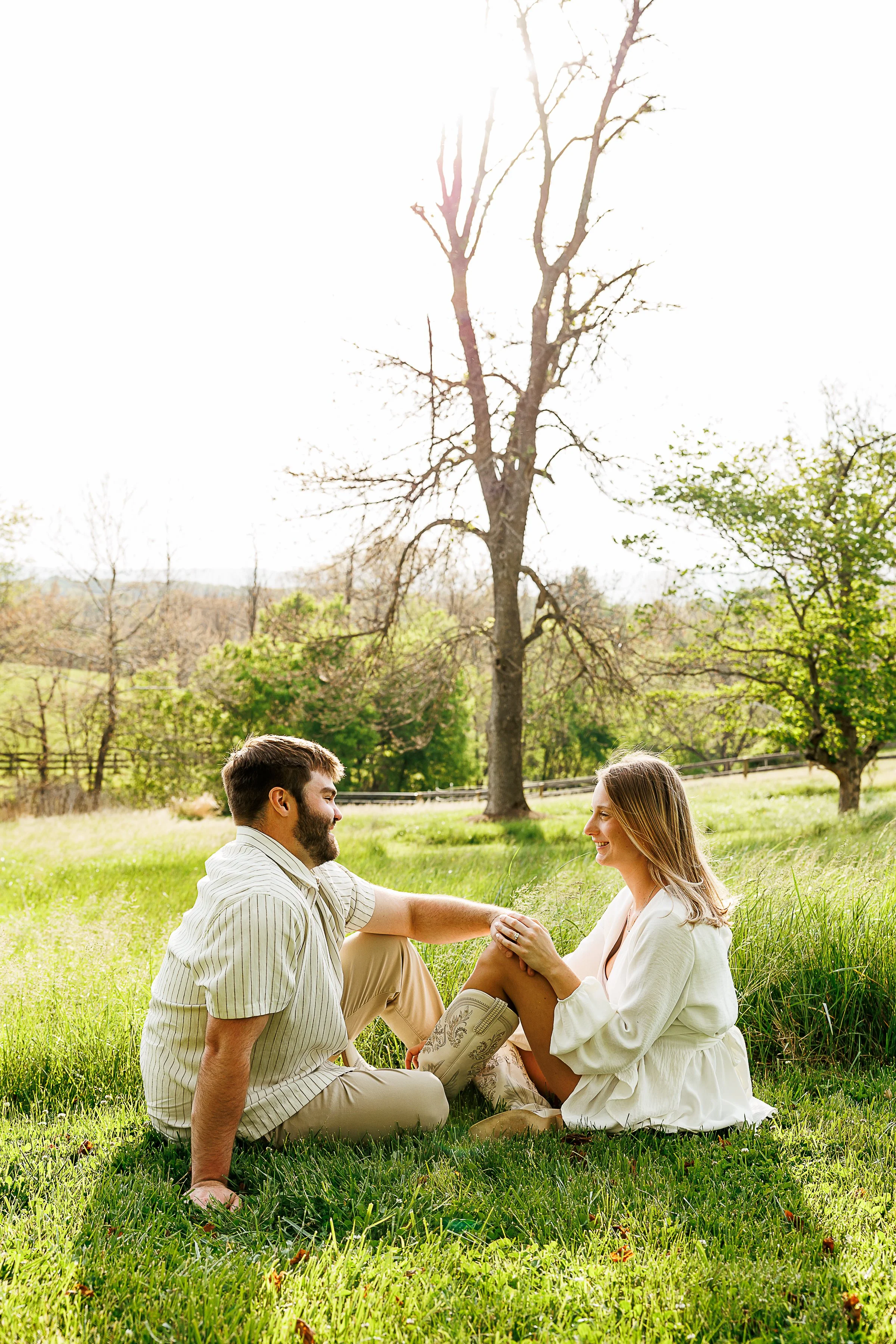 A couple sits facing each other in a sunlit grassy field, smiling as they hold hands. One partner rests an arm on a bent knee while the other wears a flowing white outfit and embroidered cowboy boots. Tall trees and bright afternoon light create a pe