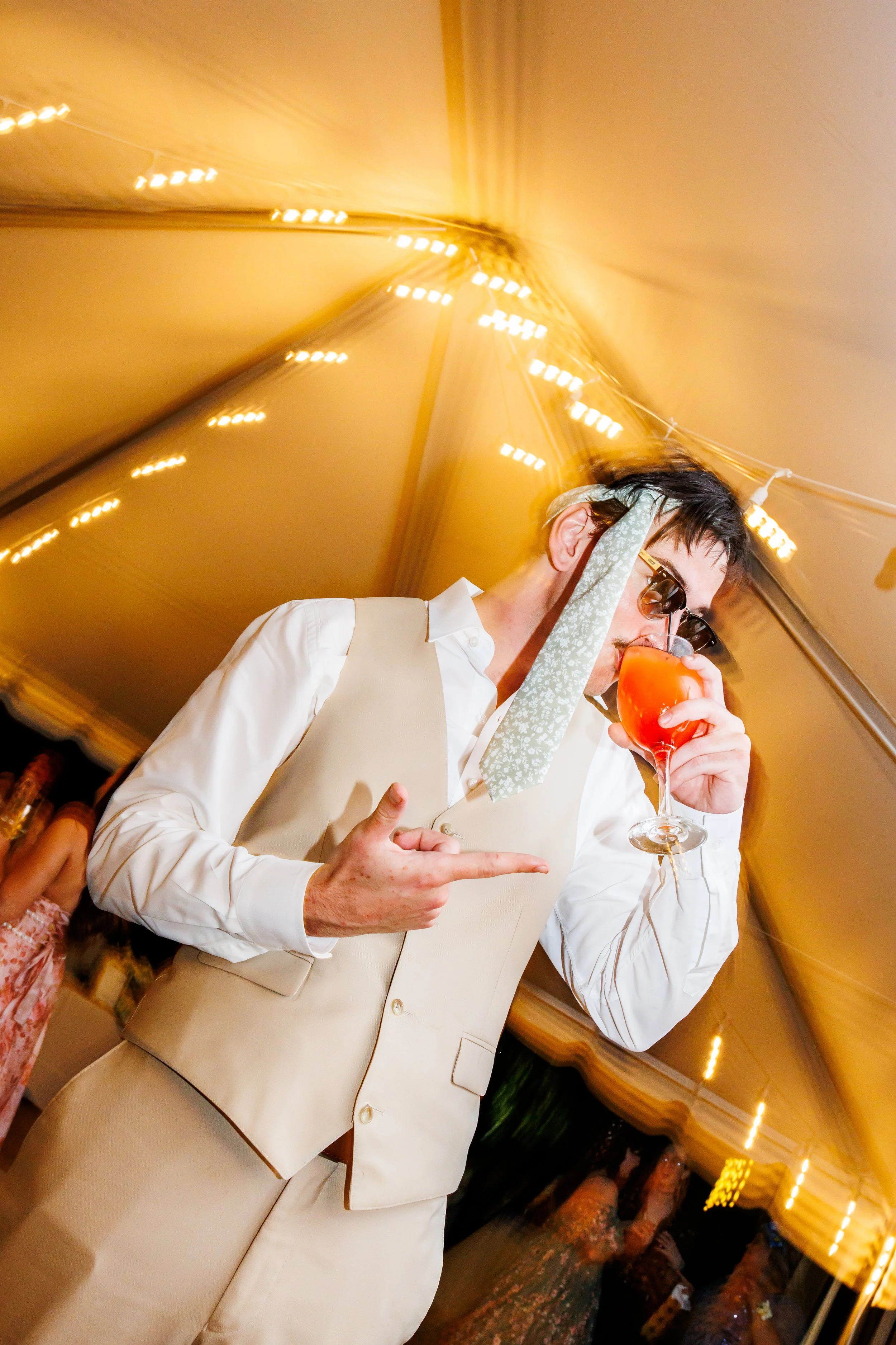 candid wedding reception portrait of a groom in a cream vest and white dress shirt dancing under a glowing tent with string lights. he wears sunglasses and a floral neck scarf while holding a bright orange cocktail and pointing playfully toward the c