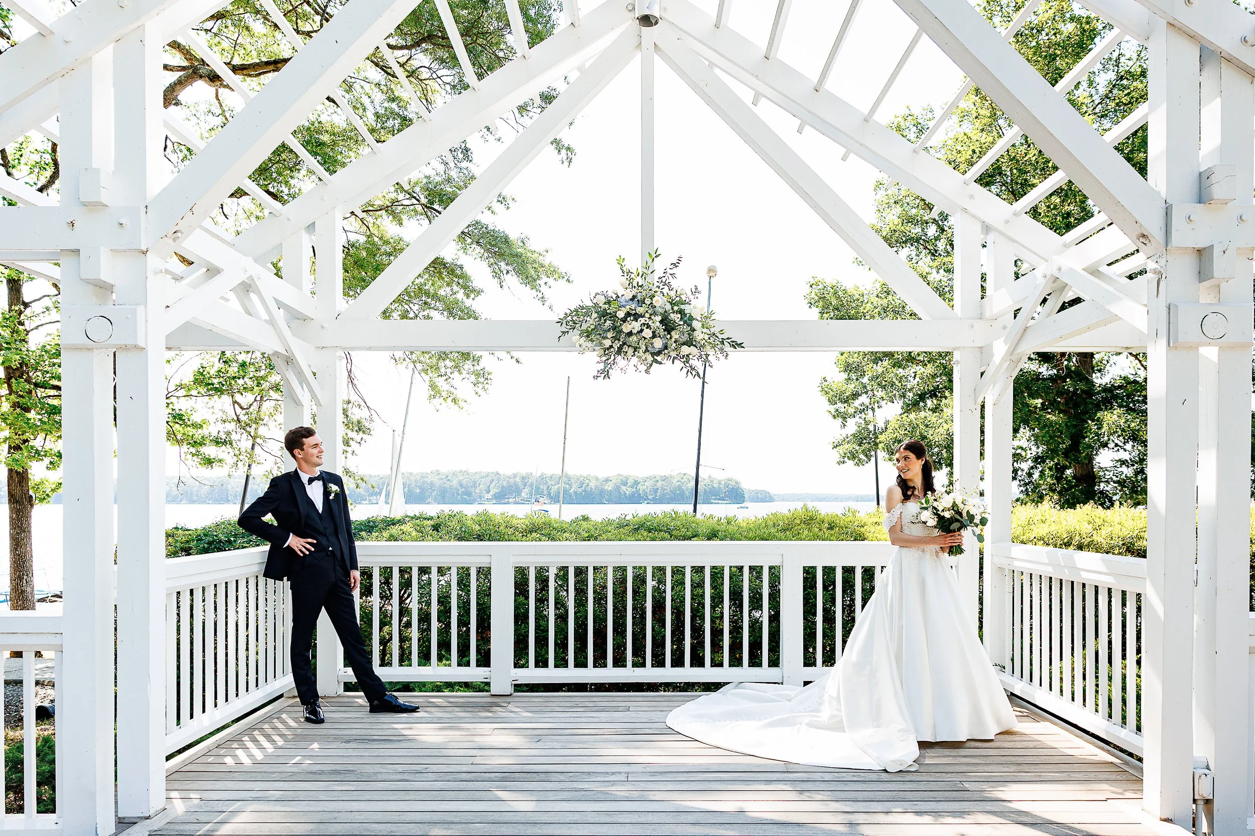 wide wedding portrait of a bride and groom standing beneath a large white open-air pavilion overlooking the water. the groom, wearing a dark suit and bow tie, leans casually against the railing while smiling at the bride. the bride wears an off-the-s