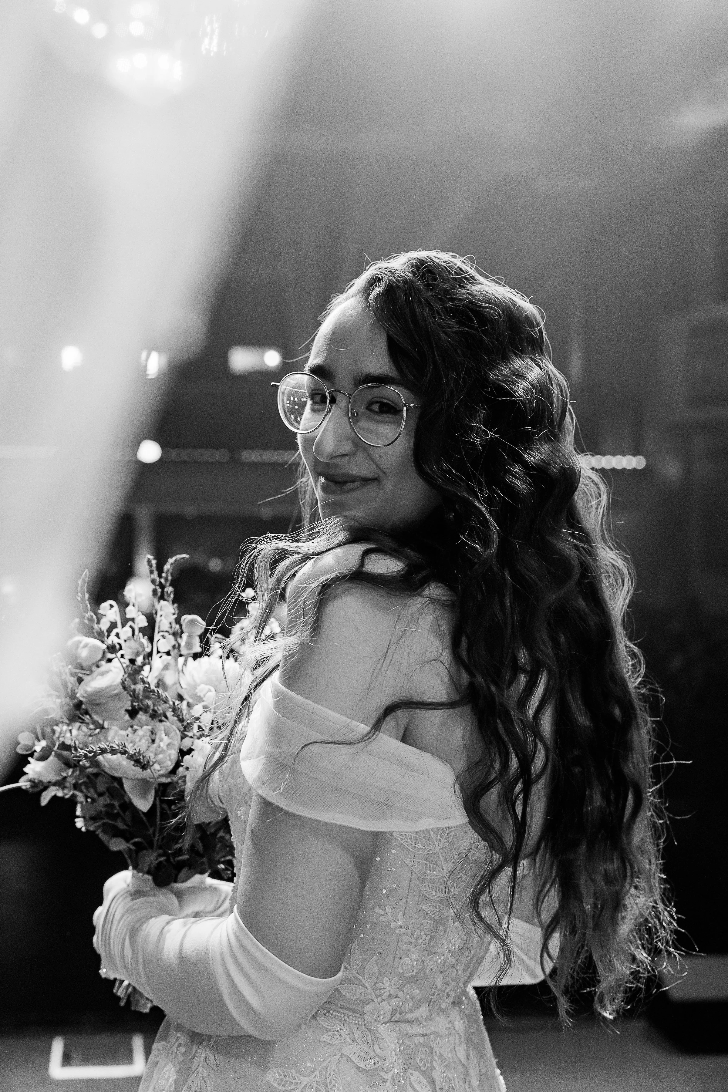 A striking black-and-white portrait of the bride as she glances back over her shoulder with a soft, confident smile. She holds her bouquet close, her off-the-shoulder gown and gloves adding an elegant, timeless feel, while light spills across her fac