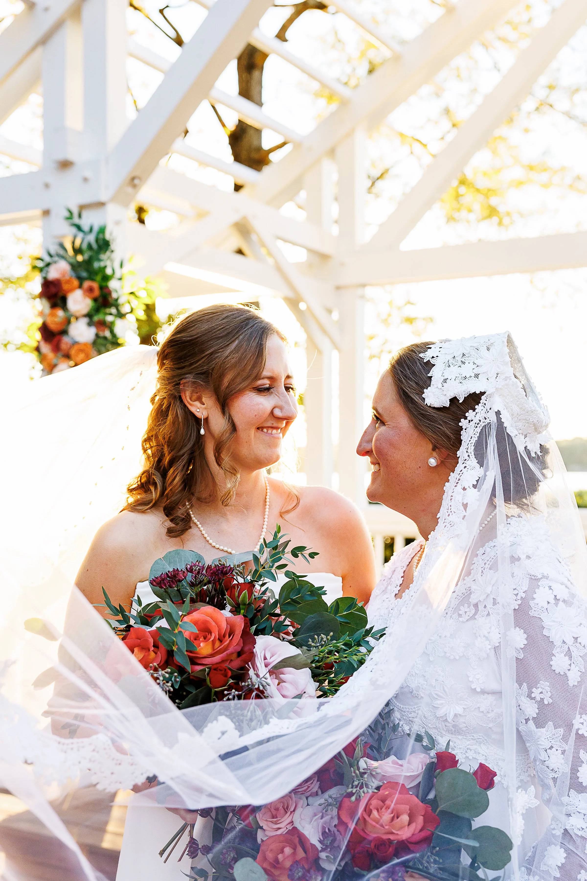 Two brides embracing under a white pergola at the Boathouse at Sunday Park in Richmond, VA, holding bouquets of red anTwo brides embracing under a white pergola at the Boathouse at Sunday Park in Richmond, VA, holding bouquets of red and orange roses