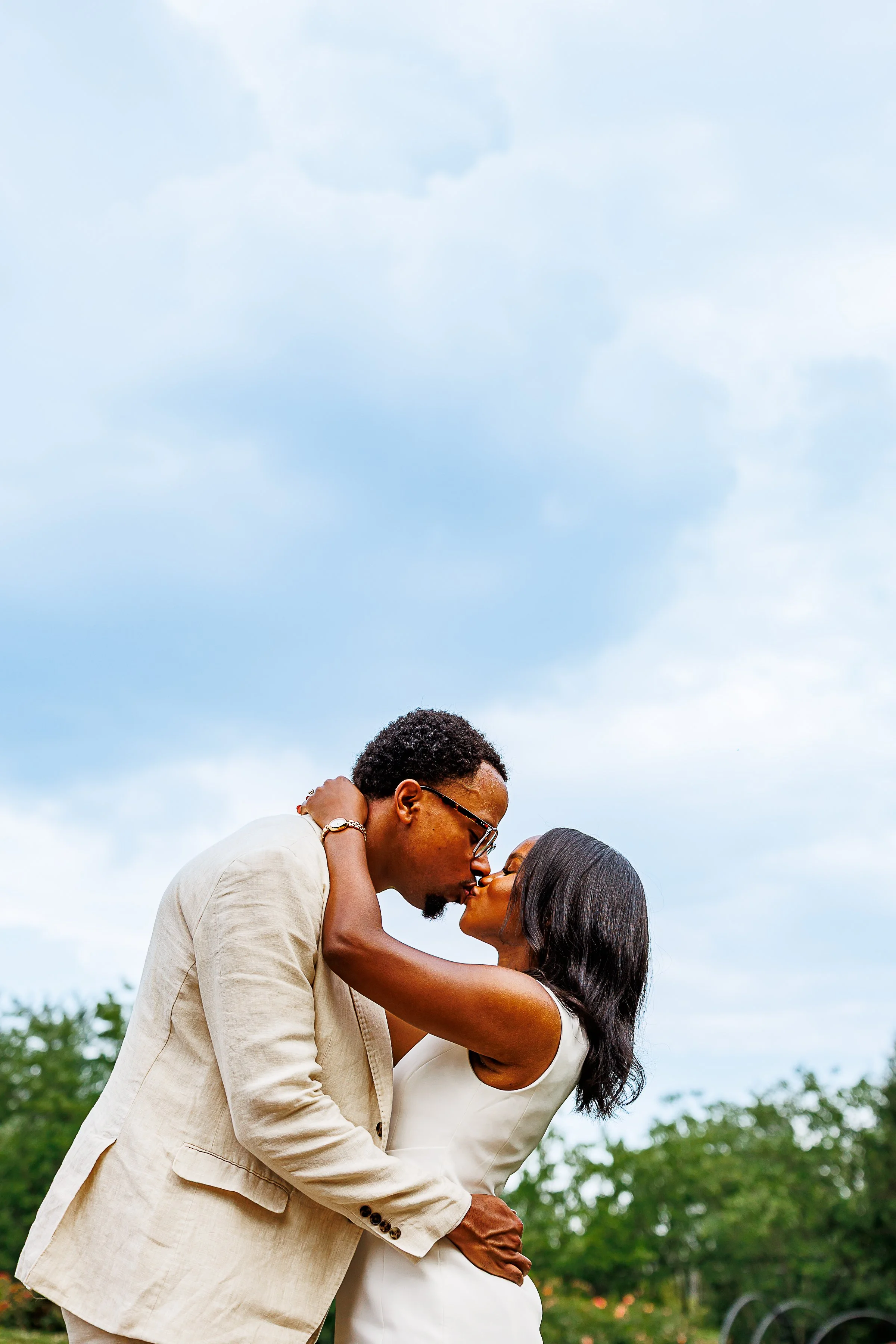 Couple embracing and sharing a kiss, him in a cream linen blazer and glasses, her in a cream sleeveless dress with soft waved hair, set against a bright blue sky with trees at Bon Air Park Rose Garden in Arlington, VA.
