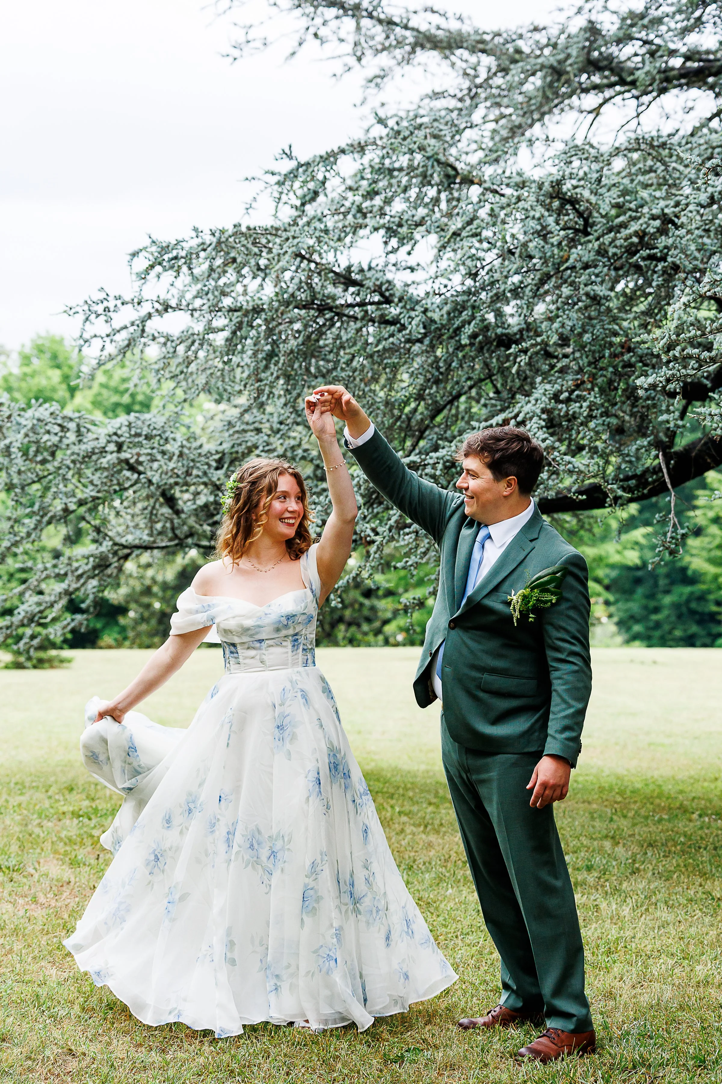 Bride and groom laughing mid-twirl under the blue cedar tree at Maymont in Richmond, VA, her floral blue-and-white gown swaying as he lifts her hand.