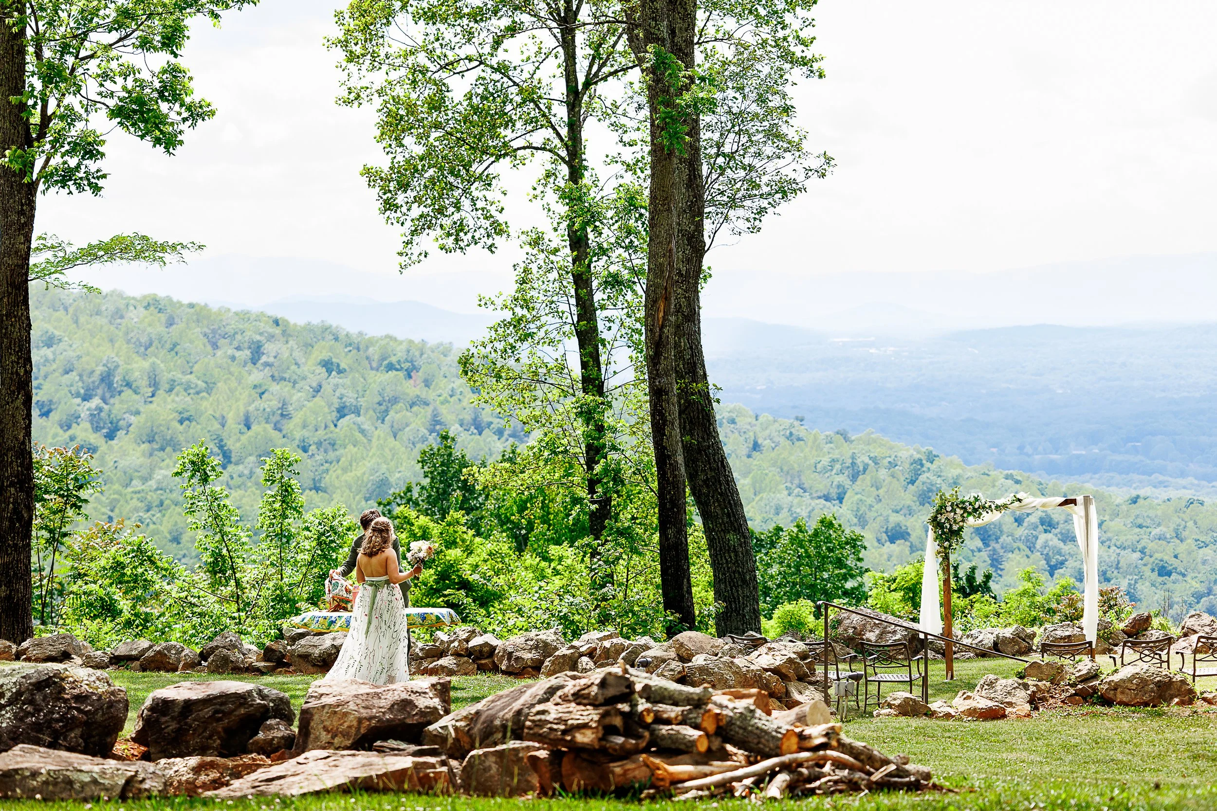 Bride and groom walk toward a floral ceremony arch on a mountaintop venue overlooking the Shenandoah Mountains near Charlottesville, Virginia, with stacked firewood and stone walls framing the scene.