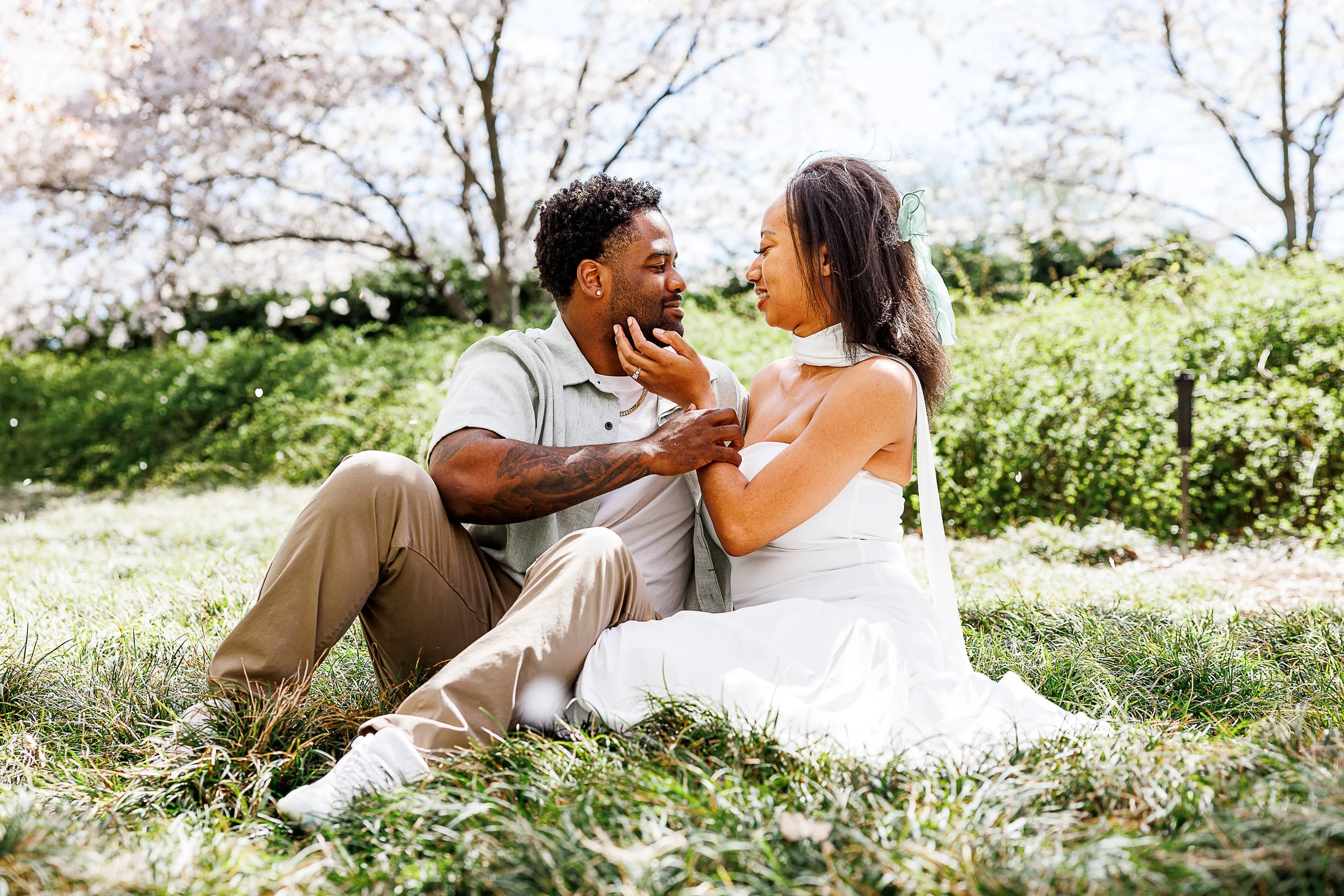 Couple sitting together in the grass during a spring engagement session in Washington, DC, smiling at each other beneath blooming cherry blossom trees.