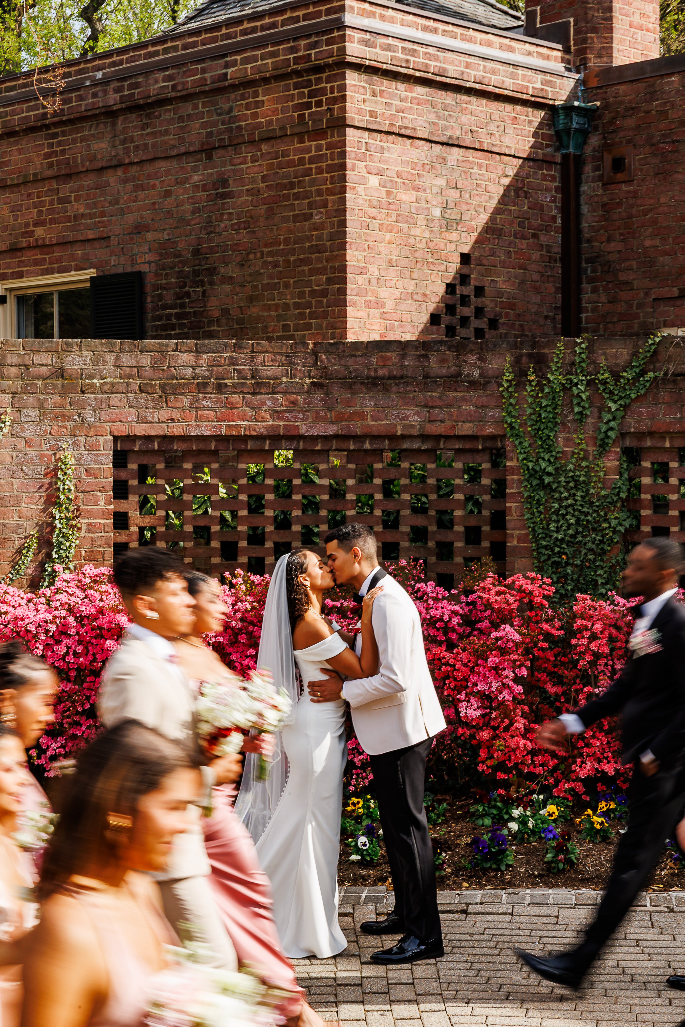 Bride and groom share a kiss in front of vibrant pink flowering bushes outside a brick venue while their wedding party walks past in the foreground, creating a dynamic motion blur. The bride wears an off-the-shoulder fitted white gown with a long vei