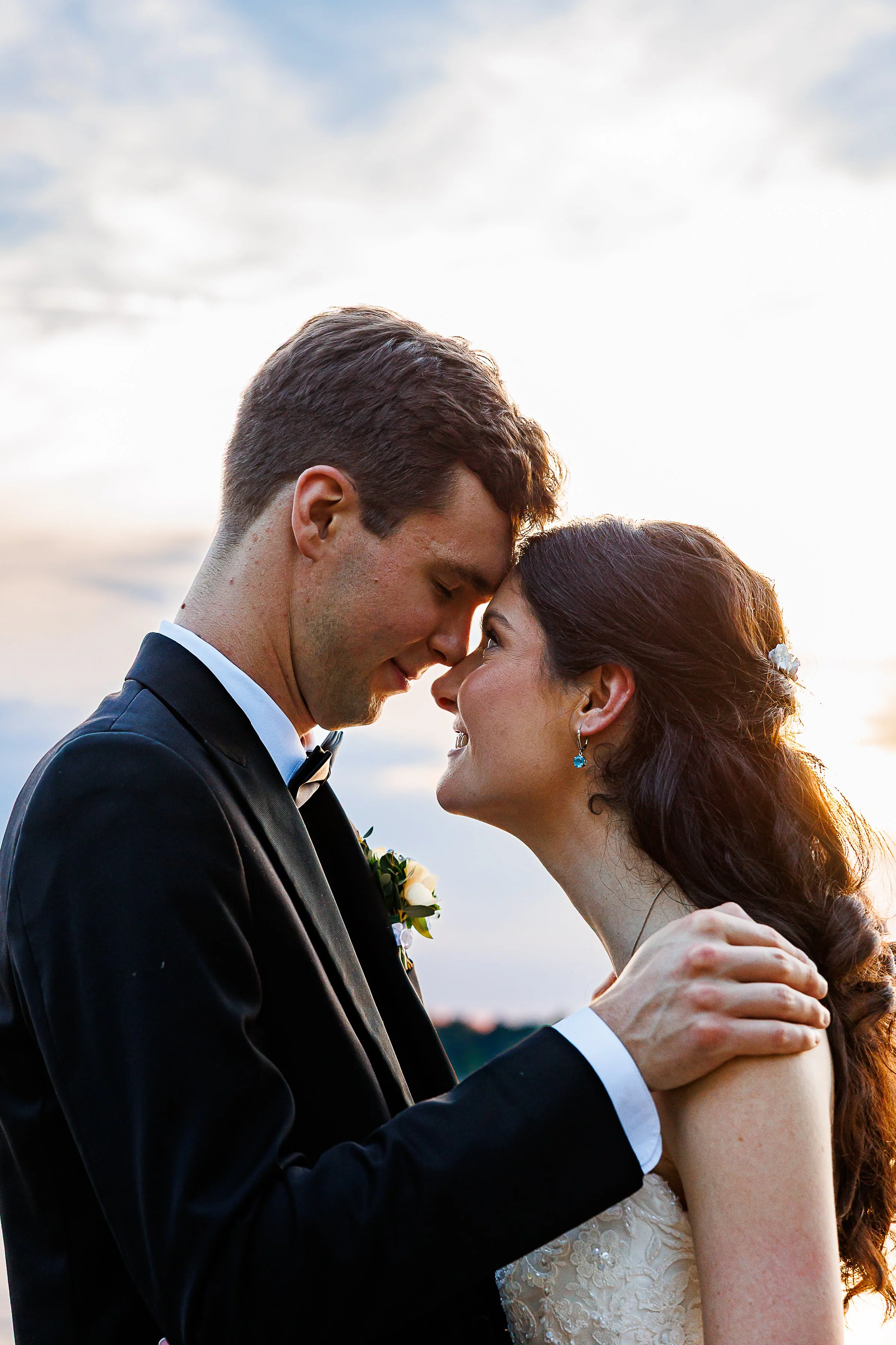 close-up sunset wedding portrait of a bride and groom gently touching foreheads and smiling at each other. the groom wears a classic black tuxedo and bow tie, while the bride wears a lace wedding gown with her dark hair styled half-up with delicate f