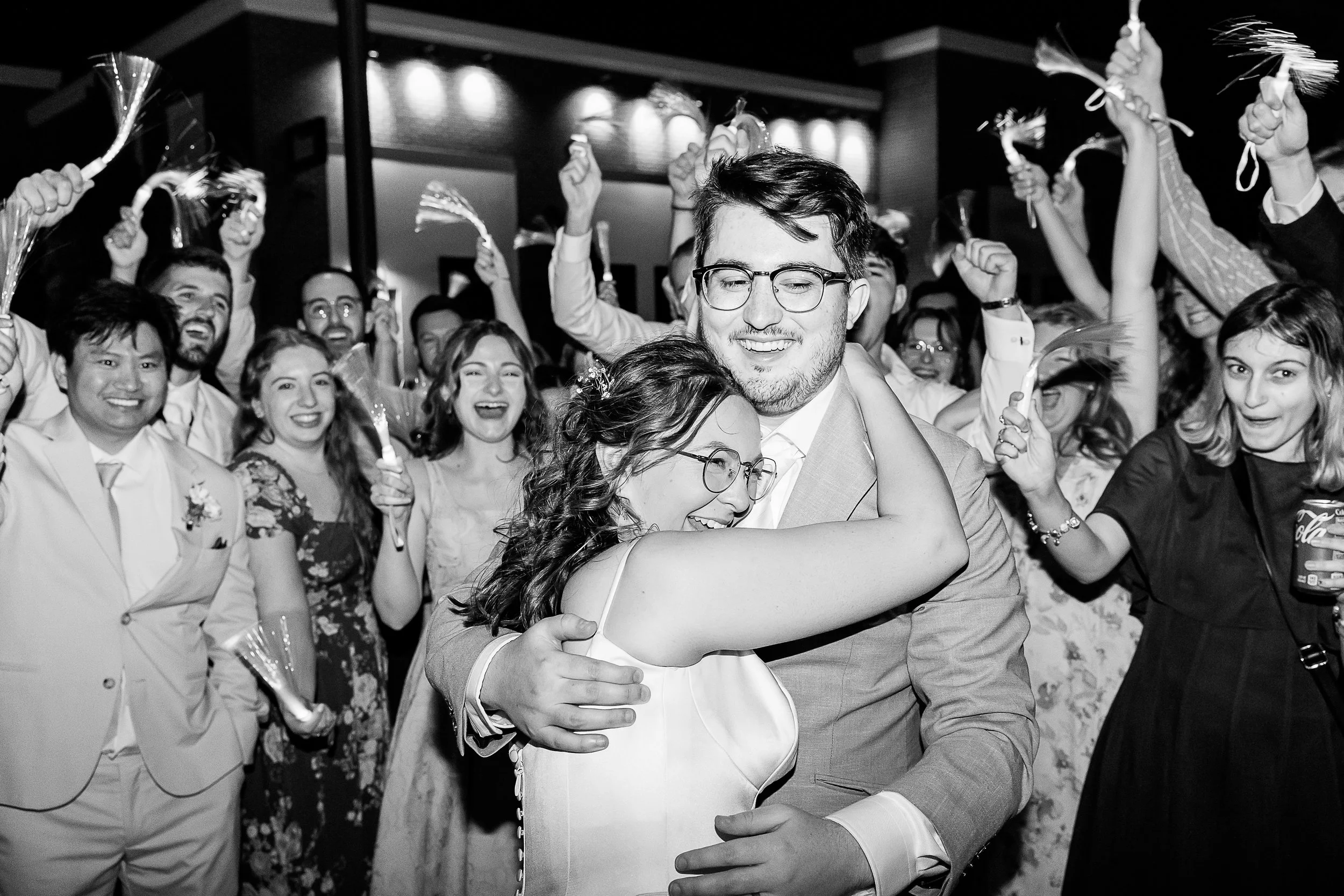 Bride and groom hugging and laughing during a lively nighttime wedding exit surrounded by cheering guests holding glowing ribbon wands.
