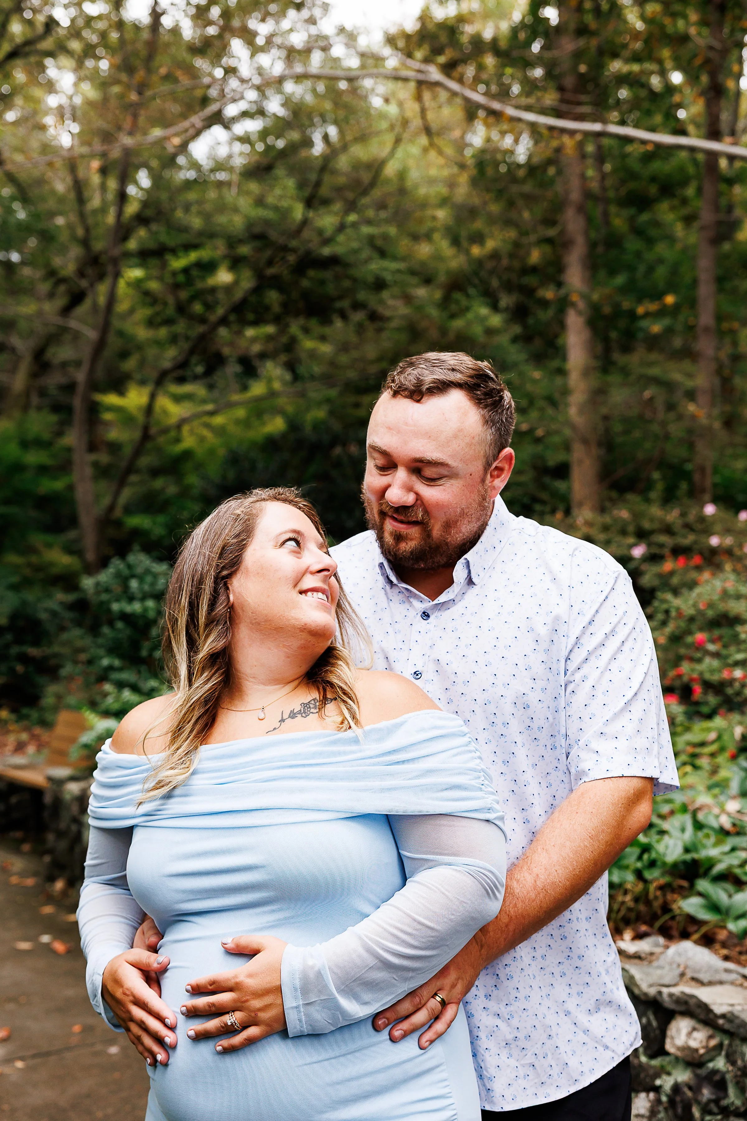 Pregnant woman in a light blue off-shoulder dress smiling up at her partner as he holds her belly, standing together in a wooded, leafy setting.