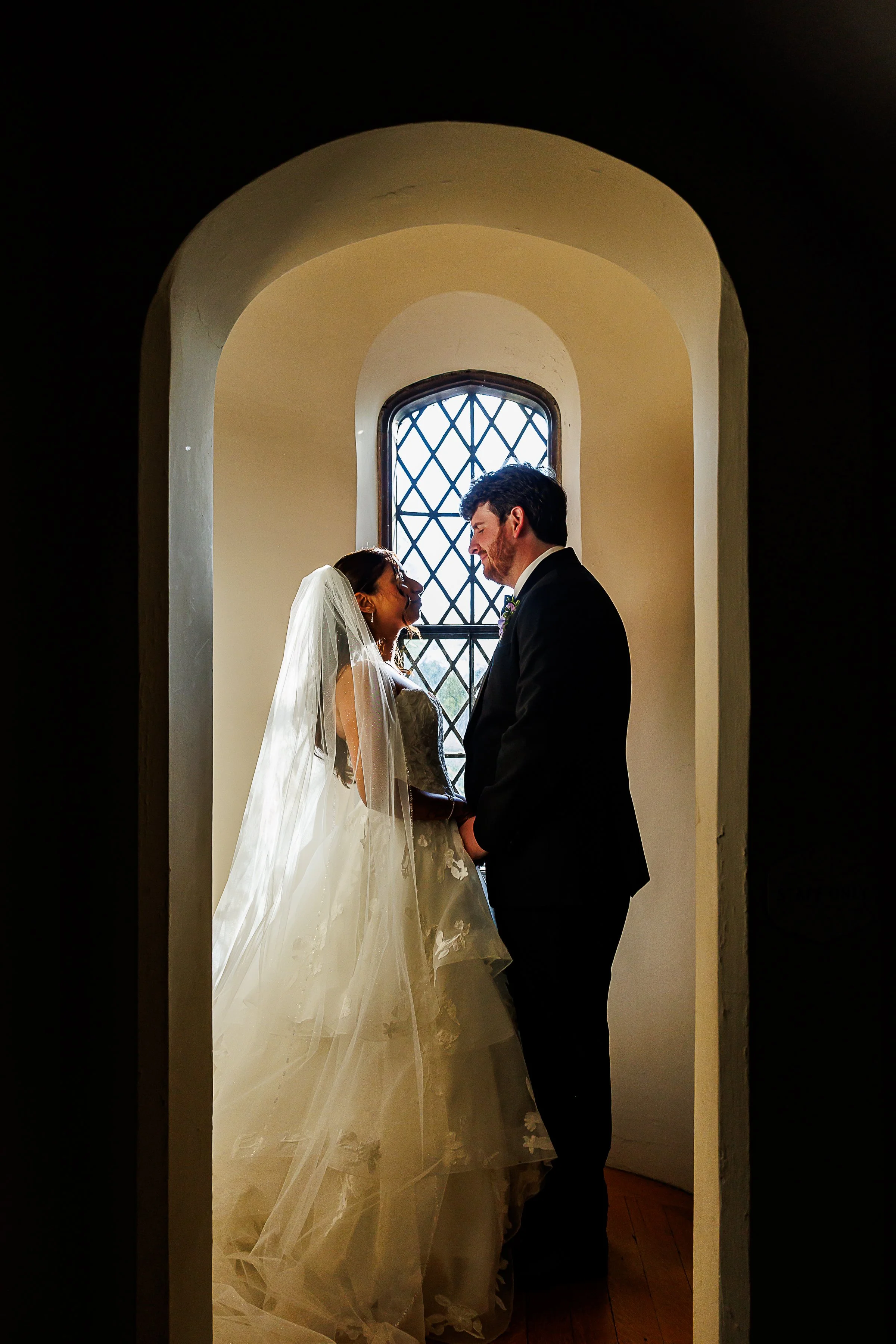 Bride and groom stand hand in hand beneath a softly lit arched window inside a historic chapel, smiling at one another. The bride's embroidered veil and flowing gown catch the light while the groom's dark suit creates a striking silhouette against th