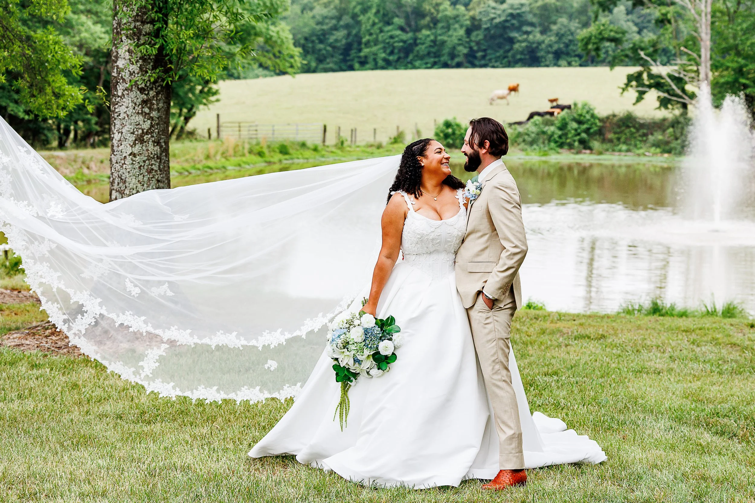 A joyful bride and groom stand together on a grassy lawn beside a pond, smiling warmly at one another on their wedding day. The bride wears a classic white gown and holds a bouquet of white and blue flowers, while her long lace-trimmed veil billows d