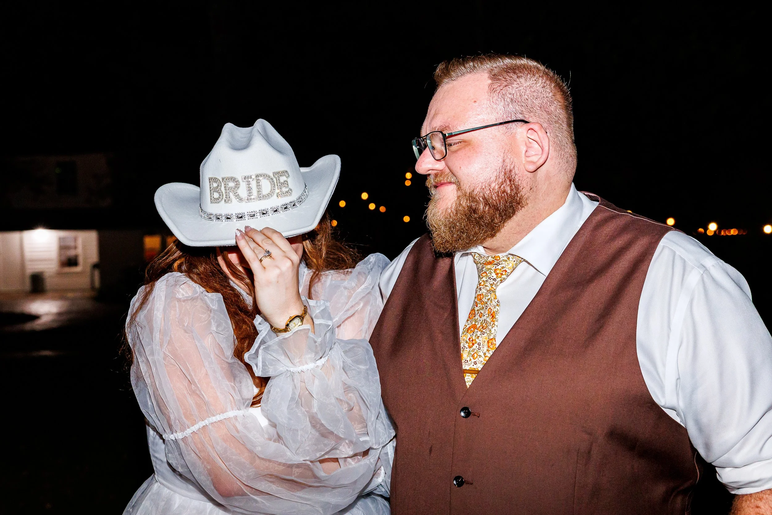 A bride wearing a white dress and sheer sleeves playfully tips down a white cowboy hat labeled “bride,” partially covering her face, while standing next to her partner at night. Her partner, wearing glasses, a white shirt, brown vest, and patterned t