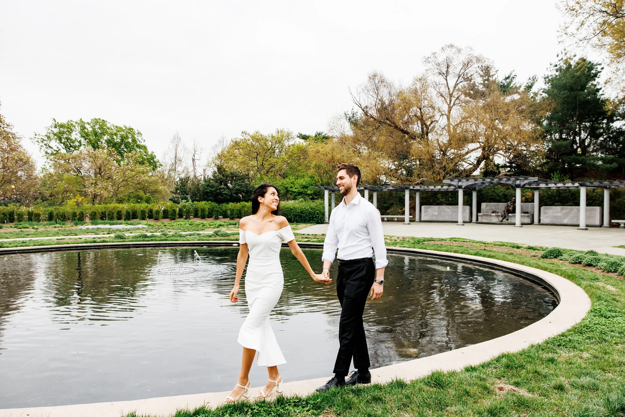 elegant engagement portrait of a couple walking hand-in-hand beside a circular reflecting pool in a formal garden. the woman wears an off-the-shoulder white midi dress and heels, while the man wears a white button-down shirt with black pants. soft gr